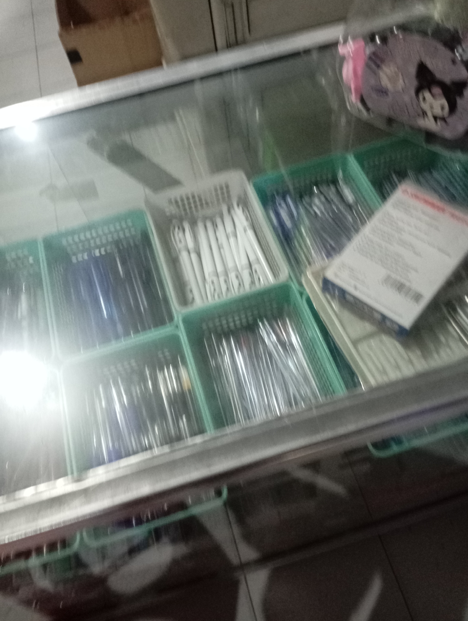 A retail display case contains several green plastic bins filled with pens. Some bins hold pens with blue ink, others with white casings. One bin contains pens with black ink. In the background, a small, purple pouch with cartoon characters is visible, along with a white box. The display case is made of glass and metal, and sits on a tiled floor. The lighting in the scene is bright and somewhat diffused, creating reflections on the glass surface. The image appears to be taken from a high angle, looking down into the display case. It is unclear if the image is from Sliyeg, Indonesia, or if it depicts any specific activity.