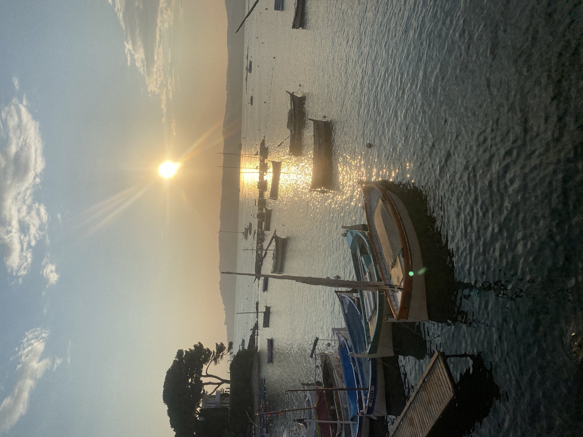 The image depicts a serene harbor scene at sunset in Antibes, France. Several small wooden boats, some with masts, are moored on the calm water. The sun, low in the sky, casts a warm golden glow on the water, creating shimmering reflections. In the background, a tree-covered hill with a building is visible, and the sky is dotted with soft clouds. The overall atmosphere is peaceful and picturesque.
