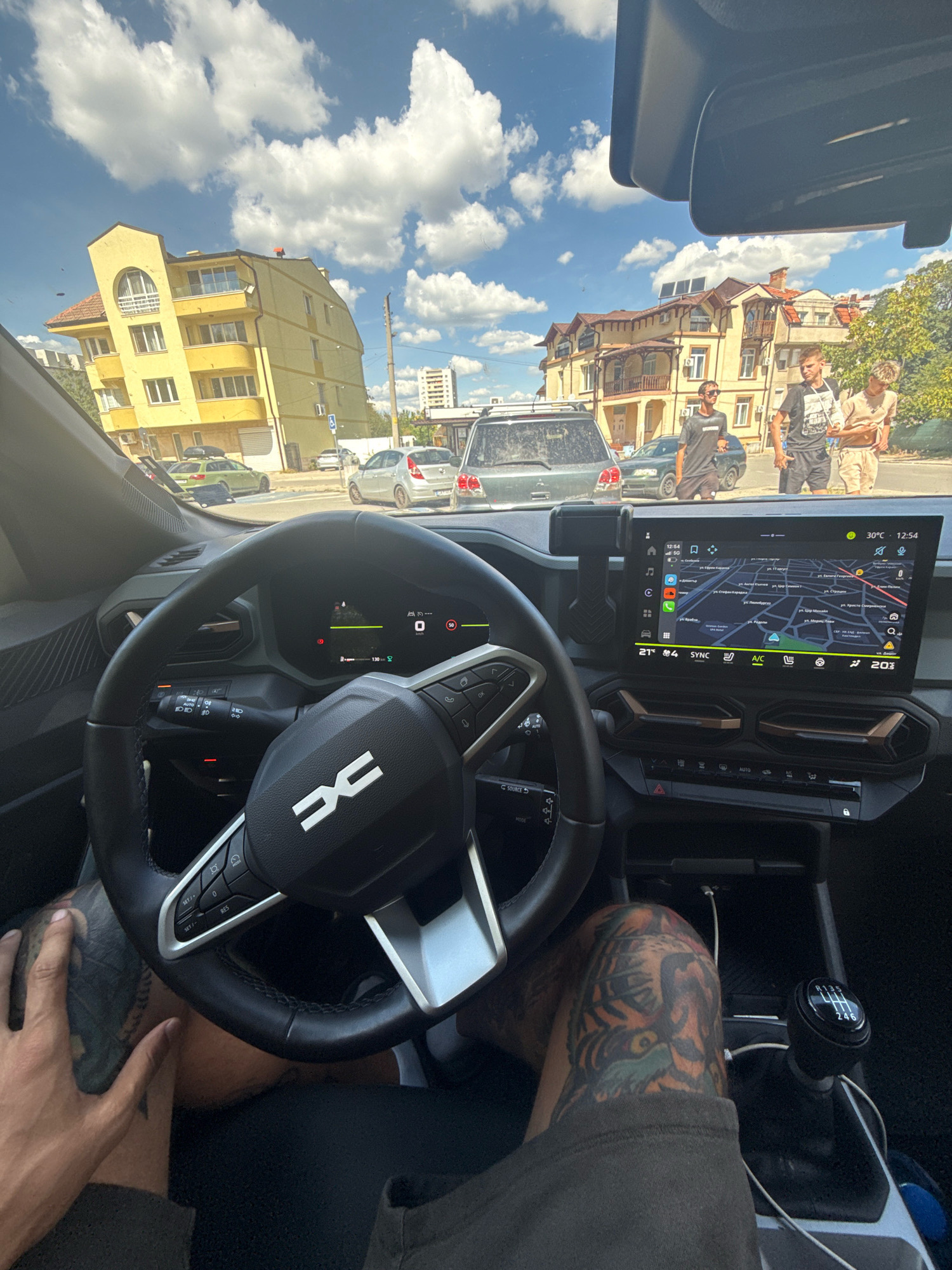 The image shows the interior of a car, presumably parked or driving in the city of Kyustendil, Bulgaria. Through the windshield, a bright, sunny day with scattered clouds is visible. The foreground is dominated by the car's steering wheel, dashboard, and a large infotainment screen displaying a map and navigation data. The driver's hands, one with tattoos, are visible resting on the steering wheel and lap.
Outside the car, several buildings are visible. A prominent yellow apartment building with a distinct architectural style stands on the left. Further down the street, a more ornate, traditional building with balconies and red-tiled roofs is present. There are several parked cars and a few people walking on the sidewalk in the background. One of the people is wearing a dark t-shirt and sunglasses, another a black t-shirt with a graphic print, and a third is wearing a light-colored t-shirt and shorts. The overall impression is a snapshot of everyday life in a European city.
The infotainment screen indicates the time is 12:54 PM and the temperature is 30°C. The navigation system shows a map of what appears to be a city in Bulgaria, with street names in Cyrillic script. The dashboard indicates the car is traveling at 50 km/h. The gear shift is visible, suggesting a manual transmission. The steering wheel has controls for audio, cruise control, and other vehicle functions. The Dacia logo is prominently displayed on the steering wheel. The presence of multiple Dacia vehicles in the vicinity could indicate the brand's popularity in this region.