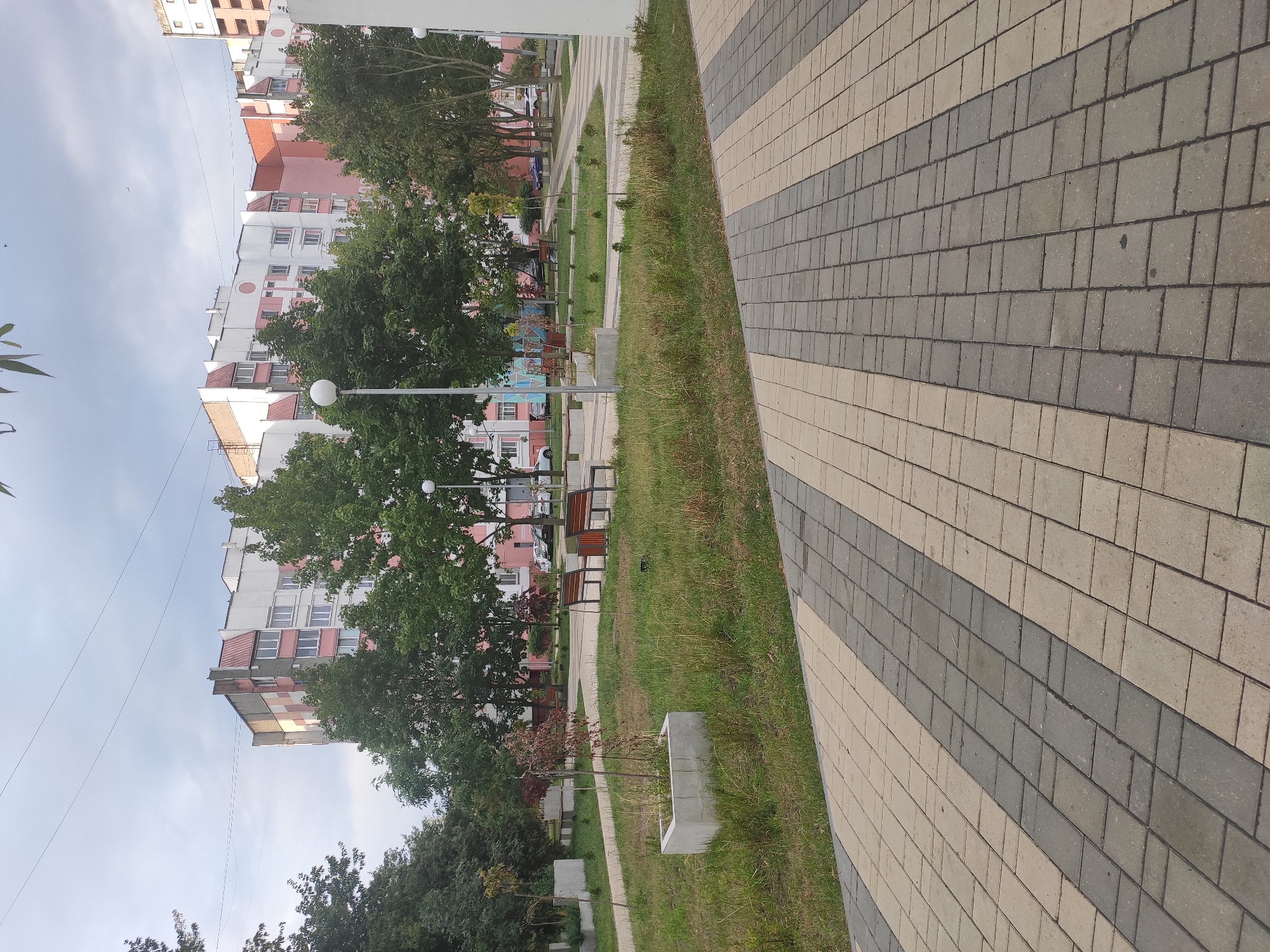 The image depicts a paved walkway with a pattern of light and dark grey rectangular bricks, leading towards a green lawn with several rectangular concrete planters. In the background, apartment buildings with white, pink, and beige facades are visible, some featuring red angular roofs. Several trees with green foliage are scattered throughout the scene. Two wooden benches with metal frames are placed on the lawn, and a lamppost with a spherical light fixture stands nearby. A few cars are parked on the street beyond the lawn. The sky is overcast with a few power lines visible. The scene appears to be a public space or residential courtyard. This is likely located in the city of Homyel, Belarus, given the architectural style of the buildings and the general aesthetic of the urban landscape.
