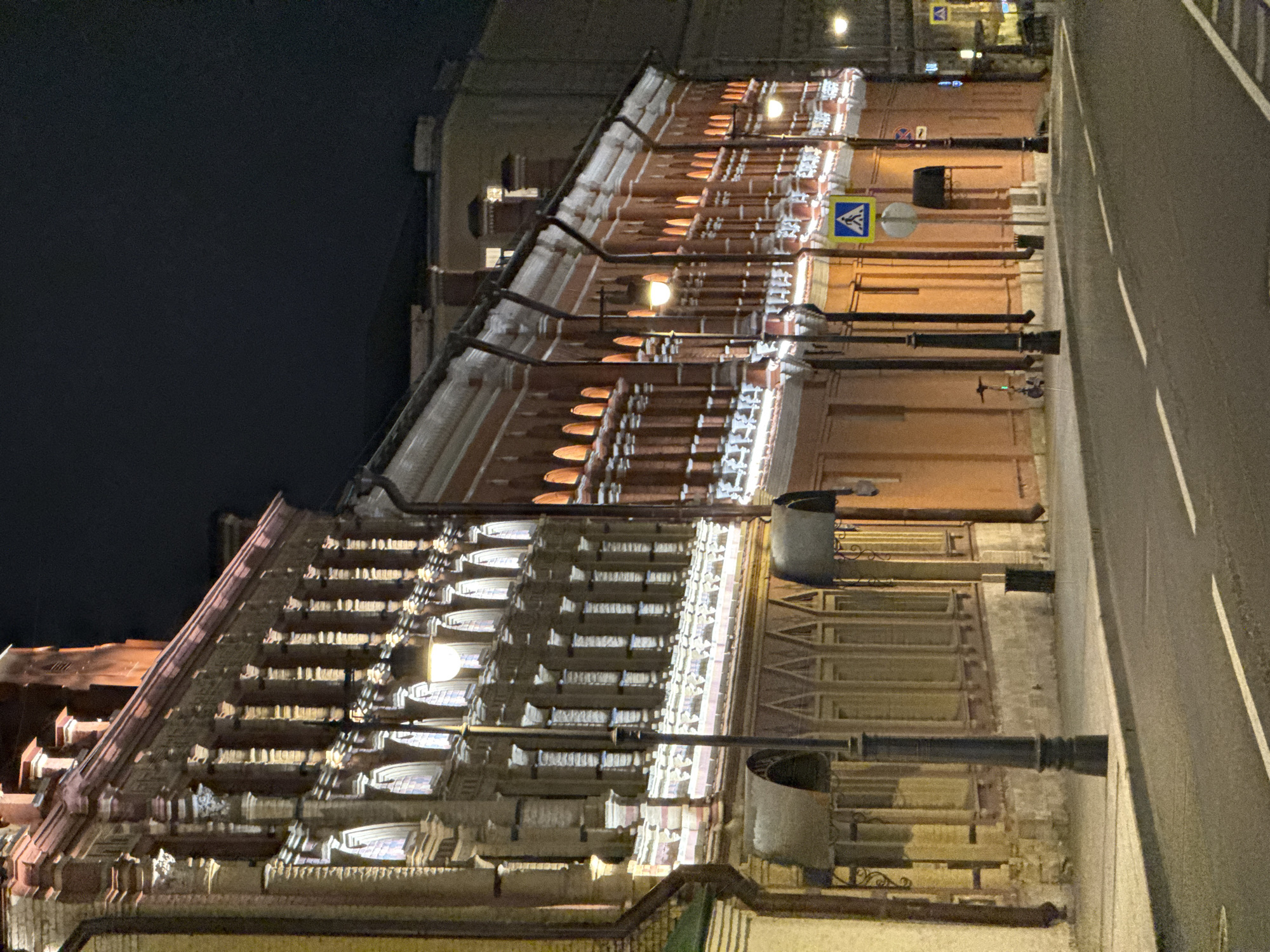 A nighttime, low-angle, full shot captures a street scene in Moscow, Russia, featuring historic brick buildings illuminated by artificial lighting. The architecture is ornate, with decorative elements like columns and arched window outlines visible along the facades. Streetlights, also lit, line the sidewalk in front of the buildings. A paved road with white lane markings dominates the foreground, reflecting the ambient light. Traffic signs, including a pedestrian crossing sign, are present. The sky is dark, indicating nighttime. No people are clearly visible in the frame. The environment appears to be an urban street with historical architecture.