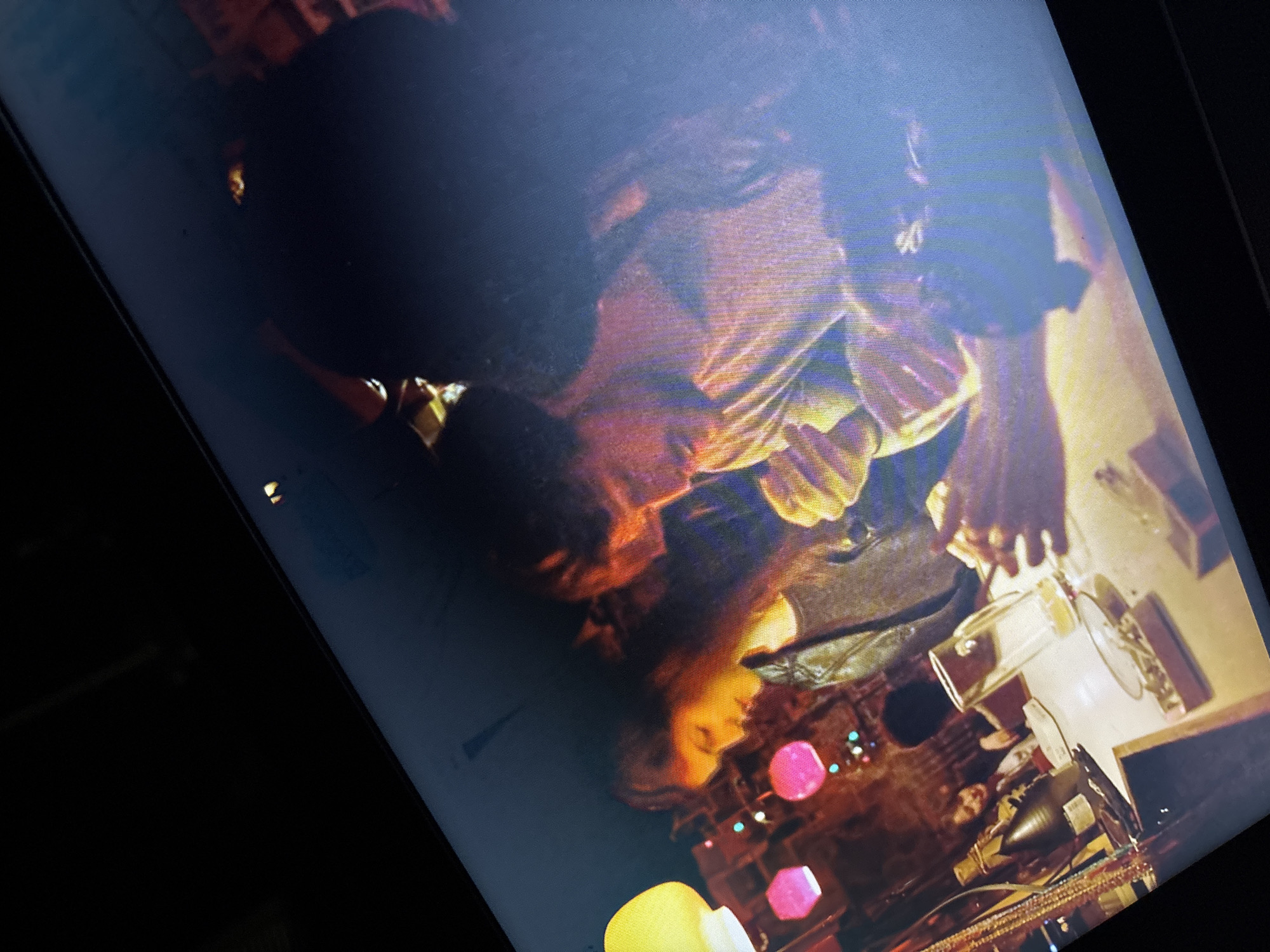 The image shows a person with long hair wearing a black jacket, a white shirt, and a necklace. They are sitting at a counter with glasses, bottles, and decorative lights. The location is Unknown, Unknown. The person appears to be interacting with items on the counter, potentially making a drink.