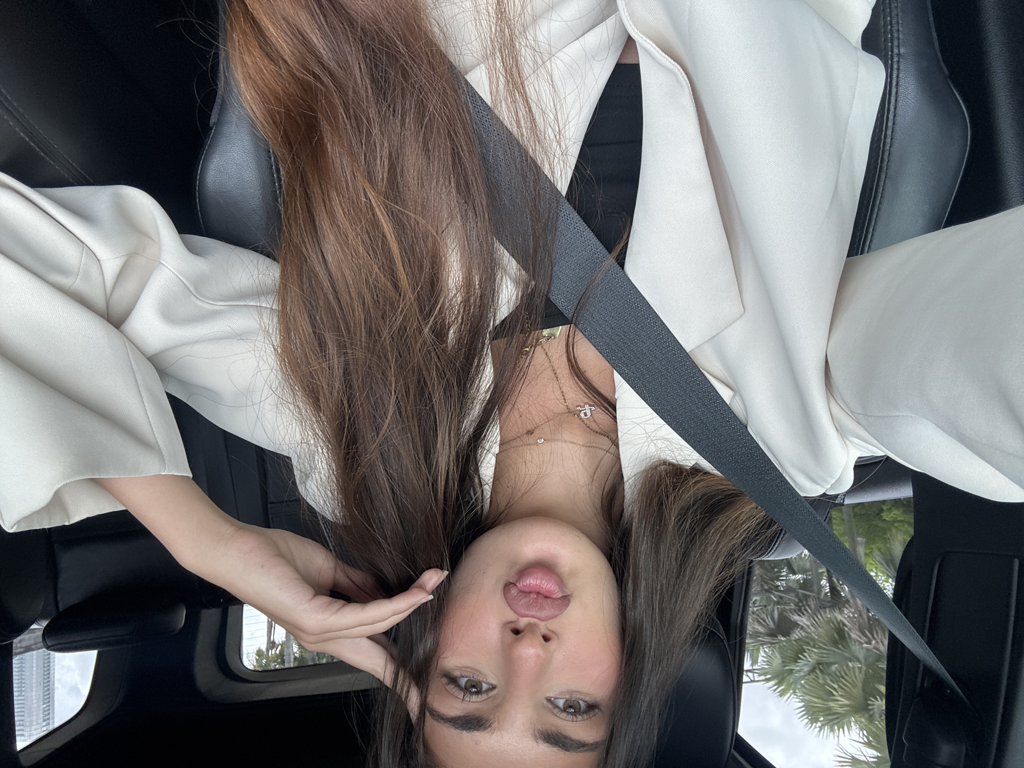 A young woman is sitting in a car, wearing a white blazer and a black top. She has long brown hair and is wearing a seatbelt. The background shows palm trees, suggesting a location in Miami, United States of America.