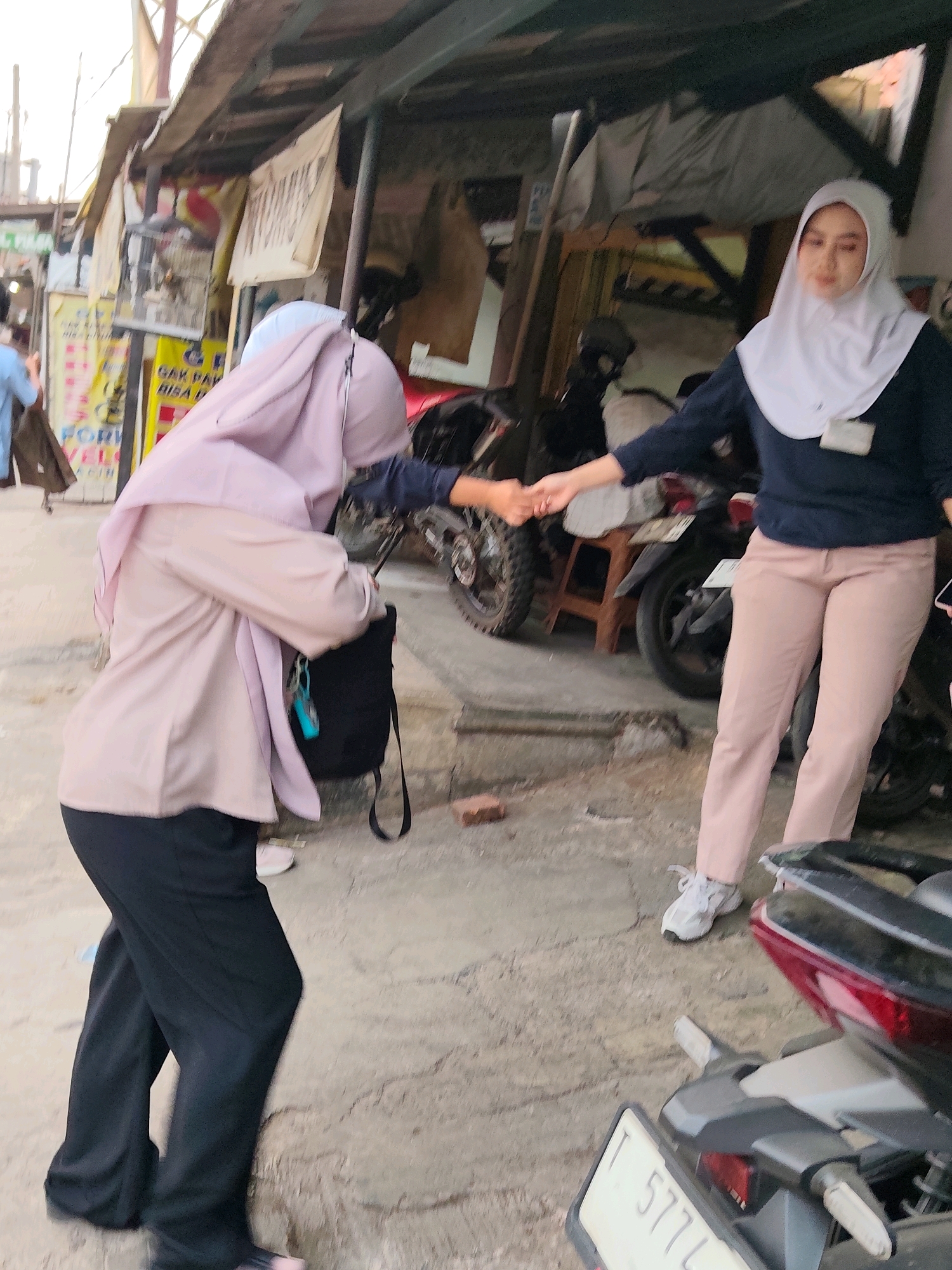 The image depicts a street scene in Purwakarta, Indonesia, characterized by a narrow alleyway lined with simple, open-fronted structures that appear to be stalls or small shops. The ground is paved with a rough, uneven surface, likely concrete or asphalt.
In the foreground, two young women, both wearing hijabs, are interacting. The woman on the left is seen from the side and back, wearing a light pink blouse and black pants, and carrying a black bag over her shoulder. The woman on the right faces the viewer more directly, wearing a white hijab and a dark blue long-sleeved top with light pink pants. They are in the process of shaking hands, suggesting a greeting or farewell.
Behind the women, several motorcycles are parked. One motorcycle in the middle ground has its license plate visible, reading "T 5774 L". The structures along the alleyway display various signs and posters, some of which are partially obscured. One prominent yellow sign has text that is difficult to decipher, but appears to be in Indonesian. There are also birdcages visible hanging from one of the stalls. The overall atmosphere is that of a busy, informal urban environment, typical of a market street in Indonesia. The lighting suggests it is daytime.
