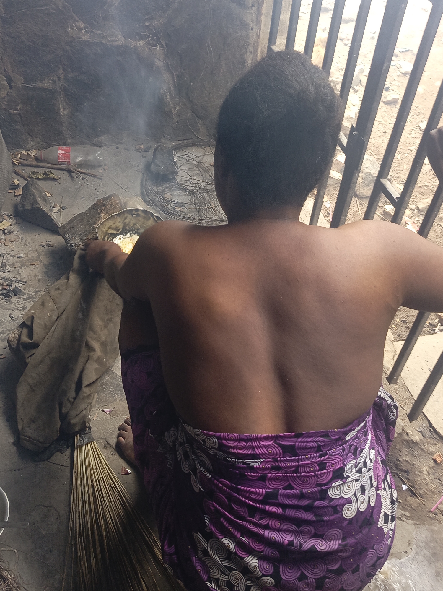 An overhead shot from behind captures a person squatting on the ground, engaged in an outdoor cooking or heating activity. The individual has dark skin and short, dark, textured hair, and is wearing a vibrant purple, black, and white patterned fabric wrapper around their lower body, leaving their upper back bare.
In front of the person, a metallic pot or bowl contains a yellowish substance, with visible smoke rising from underneath, indicating a heat source. The person's hands are obscured by the pot, suggesting they are tending to its contents. Next to the pot is a crumpled, dirty, light brown cloth or sack. To the left, a bundle of thin wooden sticks, resembling a broom, rests on the ground.
The ground surface is rough and uneven, appearing to be dirt or packed earth mixed with small stones and debris. In the background, there's a dark, rock-like wall or structure. To the right, a series of vertical metal bars, possibly part of a gate or fence, are visible, beyond which the ground continues. Among the scattered items on the ground is a clear plastic bottle with a red label, partially revealing the text "Coca-Cola." The scene appears to be taking place during the daytime in a rustic or informal outdoor setting.