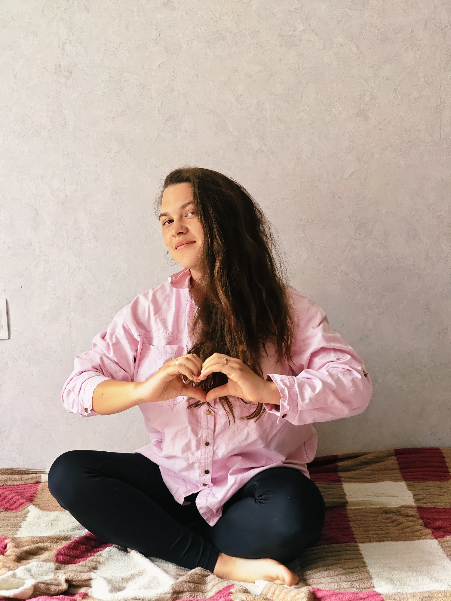 The image depicts a woman sitting cross-legged on a striped blanket, likely indoors against a light grey wall. She is wearing a pink, long-sleeved button-down shirt and black leggings. She is making a heart shape with her hands. The photo appears to be taken in Chaikovsky, Russia.