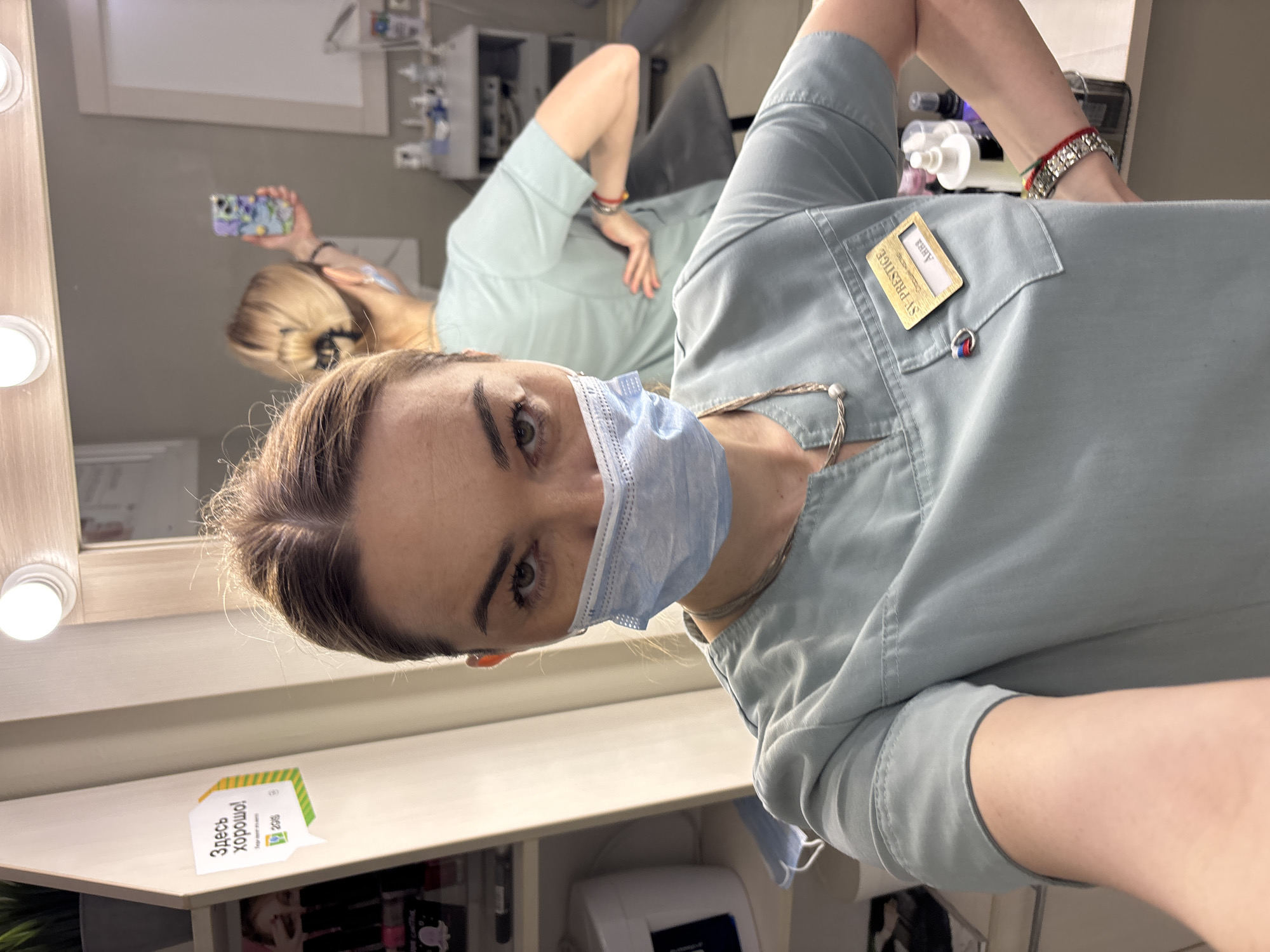 A woman in a light blue medical scrub suit and a blue medical face mask is standing in a medical facility in an unknown location. The woman is facing the camera and appears to be taking a selfie in a mirror. Reflected in the mirror is the same woman holding a phone with a patterned case. She has her blonde hair pulled back and is wearing multiple bracelets on her left wrist. On the right side of the image, attached to the scrub suit's pocket, is a golden name tag that reads "St. Prestige". The environment includes a light wood counter surface with a small sign in Russian, "Здесь хорошо" (Here is good) and what appears to be medical supplies on a shelf under the counter and in the background. The lighting suggests an indoor setting with artificial light sources.