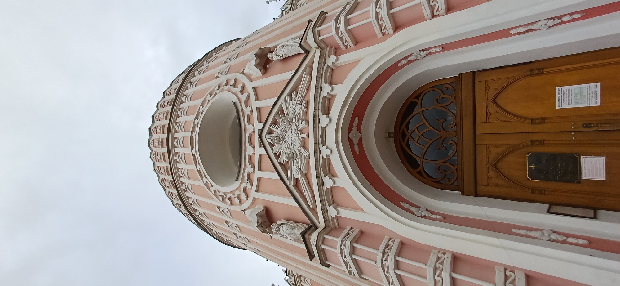 The image shows a low-angle shot of the exterior of the Church of the Nativity of the Virgin Mary, a Roman Catholic church located in Saint Petersburg, Russia. The church features a distinctive pink facade with white decorative elements. The architectural style is Neo-Gothic, evident in the pointed arches, ornate detailing, and statues. The foreground is dominated by a large wooden double door with a decorative arched window above it. Several signs are affixed to the doors, one of which features a cross. Two white statues of figures are visible on either side of the building's upper section. The sky above is overcast and grey.