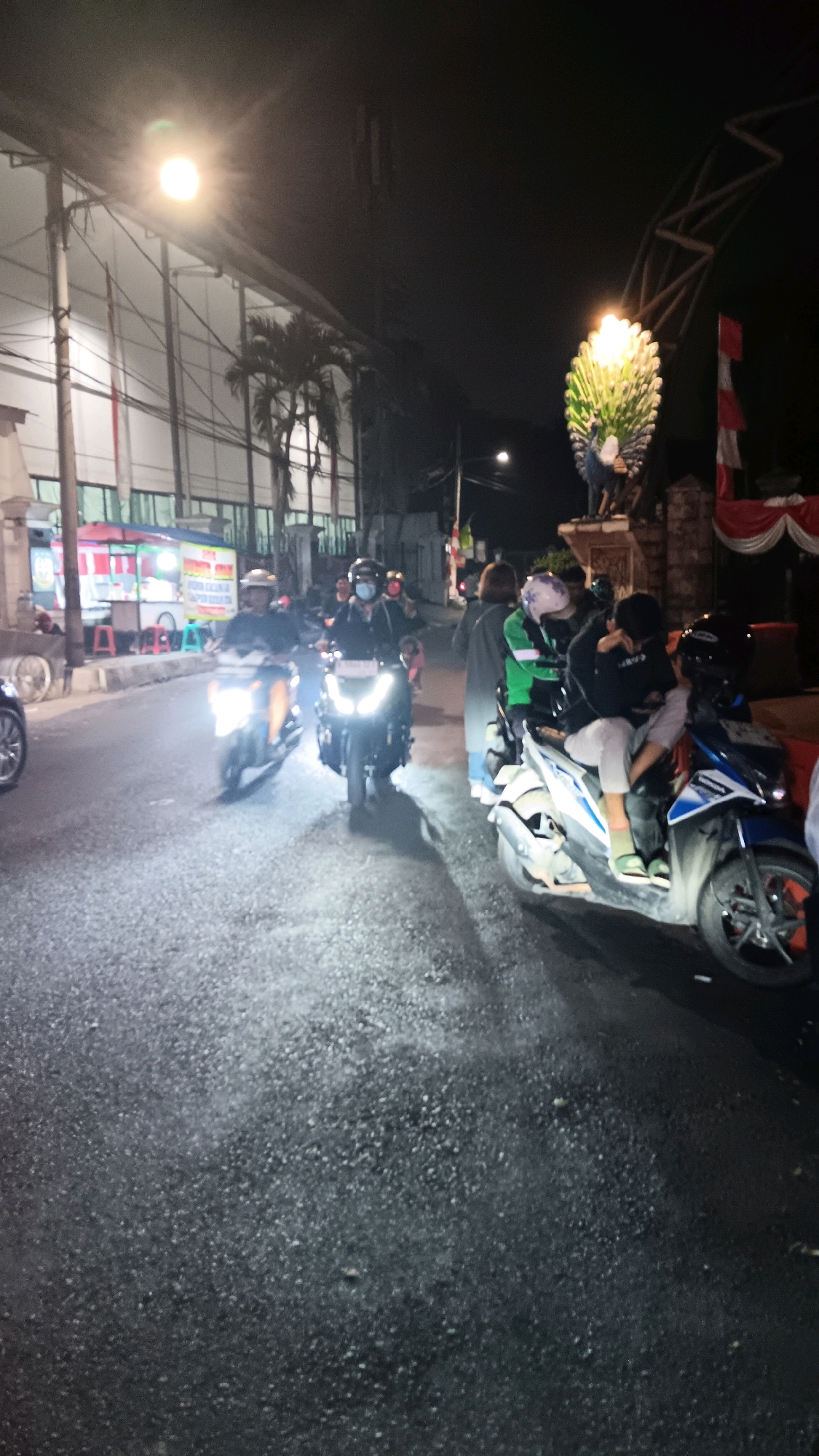 At night on a street in South Jakarta, Indonesia, three motorbikes are in motion. The motorbike closest to the viewer is blue and white, carrying two individuals. The middle motorbike is black, with a single rider. The furthest motorbike is orange and white, also with a single rider. A large, illuminated peacock statue stands on the right side of the street, near an arched gateway adorned with red and white banners. Buildings with fluorescent lights are visible on the left side of the street, with palm trees interspersed between them. The street is paved with asphalt and appears wet, reflecting the lights of the motorbikes and overhead lamps. The scene depicts typical urban nightlife activity.