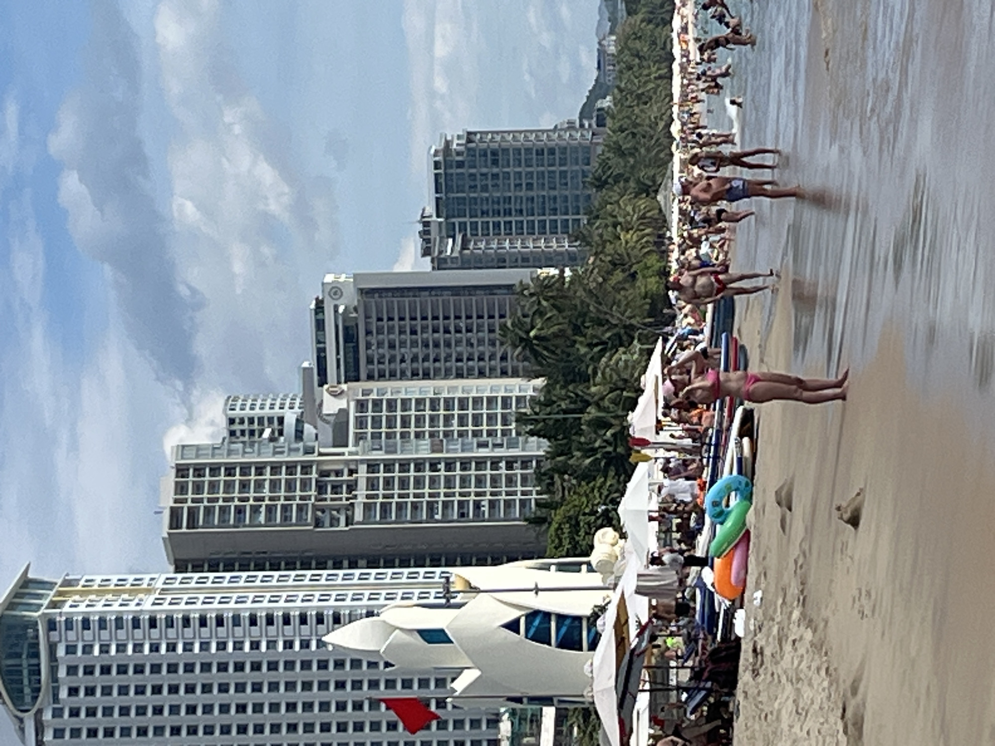 The image depicts a crowded beach scene in Nha Trang, Vietnam, with the iconic Lotus Tower prominently visible in the background. The beach is lined with numerous high-rise buildings, characteristic of the city's coastal development. A stretch of sandy beach is visible in the foreground, with several beach umbrellas and lounge chairs set up for visitors. Numerous people are scattered across the beach and in the shallow waters, engaged in various leisure activities. Some are sunbathing, while others are wading in the sea or playing in the water. Beach toys, including inflatable rings and surfboards, are scattered on the sand. The water appears relatively calm, with gentle waves lapping at the shore. The sky is partly cloudy, suggesting a bright but not intensely sunny day. The overall impression is one of a popular tourist destination bustling with activity.