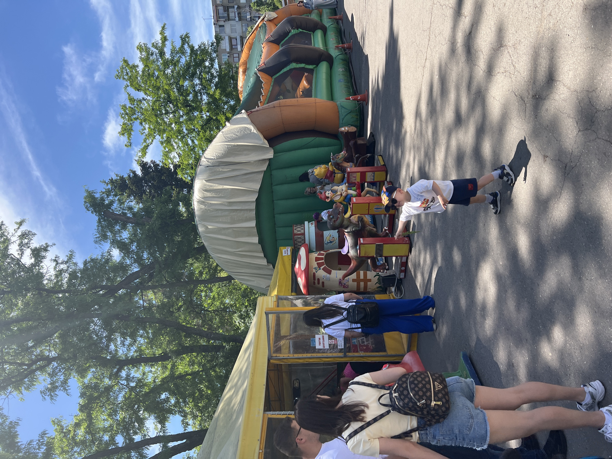 A fairground in Sofia, Bulgaria, on a sunny day features an inflatable castle, a carousel with animated animal figures, and a ticket booth. The carousel is painted with bright colors and whimsical characters. A child in a white T-shirt, blue shorts, and a baseball cap stands near the carousel. Two people stand near the ticket booth, one wearing blue pants and the other in denim shorts with a backpack. The background includes trees, a blue sky with clouds, and a multi-story building. The ground is paved and partly shaded by the trees.