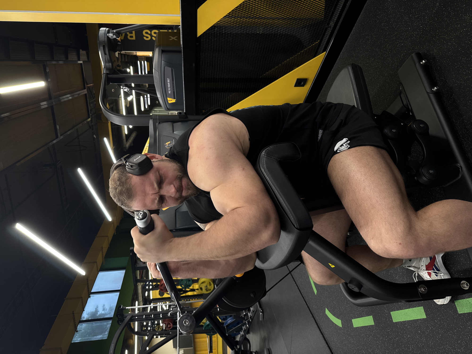 A male individual is actively engaged in a gym setting, seen from a high-angle, slightly rotated perspective. He is positioned on a black leg curl machine, appearing to be in the middle of an exercise, with his body bent forward and hands gripping the handles for stability. He wears black Marshall headphones, a black sleeveless top, black shorts, and white athletic shoes with red and blue accents. His face shows concentration and exertion, with his eyes closed or looking downwards.
The gym features a modern design with prominent black and yellow color schemes on walls and equipment. Overhead, long, bright linear LED lights illuminate the space. In the background, other gym equipment is visible, including racks with colorful weights and another machine labeled "PEC FLY / REAR DELT." Through large windows or reflective surfaces on the left side of the image, an overcast sky and outdoor trees can be glimpsed, suggesting it is daytime. The floor is dark with bright green rectangular tape markings, possibly indicating workout zones. A partial text "FITNESS RA" is visible on a yellow structure in the upper background, likely part of a gym name like "FITNESS RAGE." A yellow warning symbol is also present on the leg curl machine.