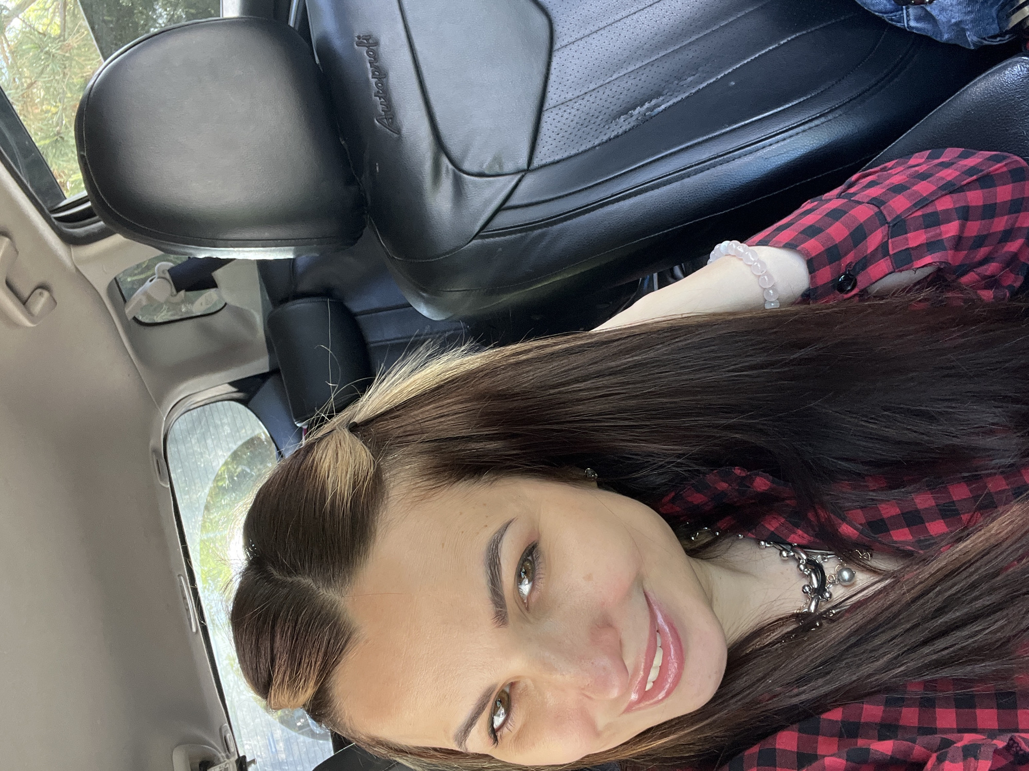 A woman is seated inside a car, wearing a red and black checkered shirt. She has brown hair pulled back at the temples and is smiling at the camera. She is wearing a silver necklace and a bracelet. The car's interior is visible, with black leather seats and a headrest bearing the word "Autoprofi." Outside the car window, blurry green foliage can be seen. The location is Unknown, Unknown.
