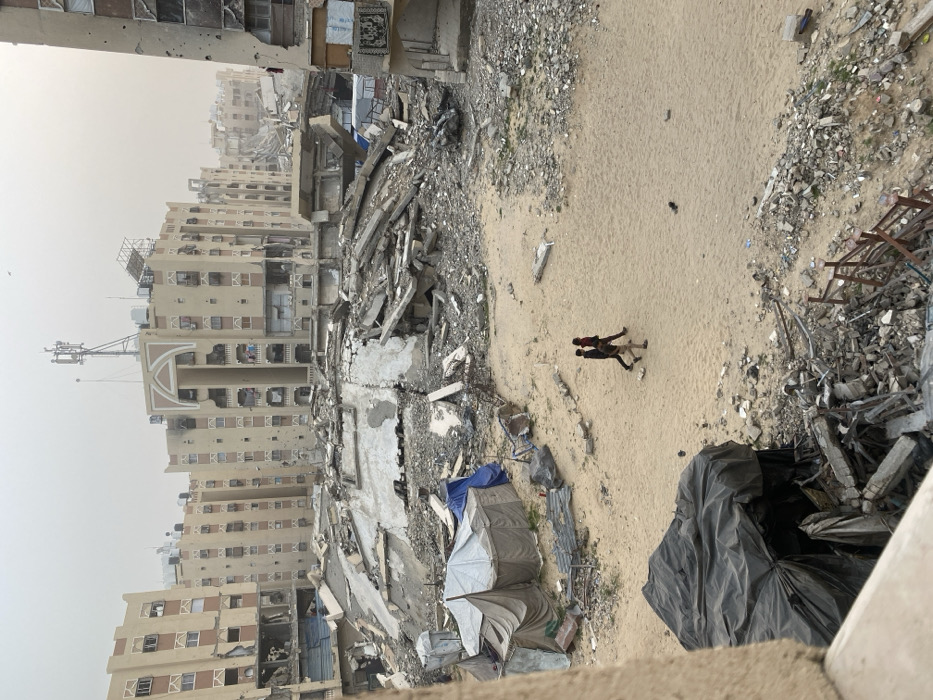 The image depicts a scene of extensive structural damage in Khan Yunis, Palestinian Territory, viewed from an elevated perspective. In the foreground, a concrete ledge is visible, with a dark tarp covering debris in the bottom left. The midground features an irregular sandy clearing, across which three individuals, likely young men, walk from left to right. To the left of the clearing, two large, light-colored canvas tents and a blue tarp are situated, forming a makeshift encampment. Surrounding the sandy area and extending into the background are massive piles of concrete rubble, twisted rebar, and debris from collapsed buildings.
In the background, multiple multi-story residential buildings, primarily light beige, exhibit severe damage, including numerous impact holes on their facades, missing wall sections, exposed interiors, and damaged balconies. One prominent building in the central background features an ornate arched entrance or balcony design and communication towers on its roof, also showing signs of damage. The sky is overcast and hazy, indicating a dull, possibly dusty, atmospheric condition. The scene conveys widespread destruction and makeshift living conditions amidst the ruins.