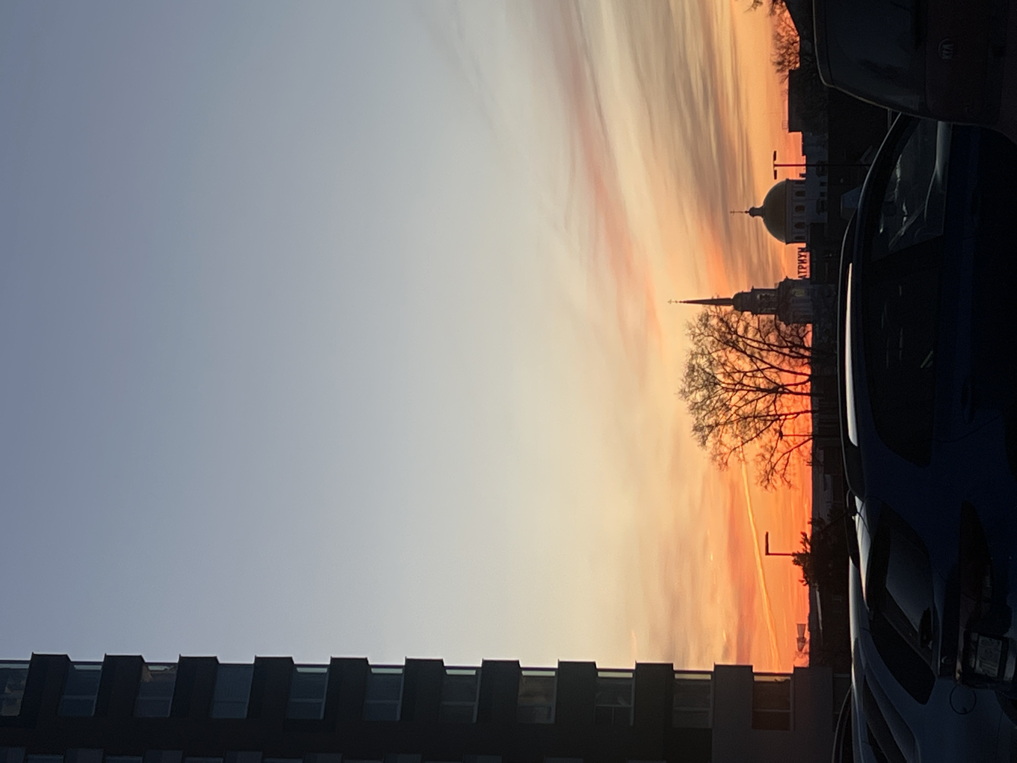 The image shows a sunset scene in what appears to be an urban setting. The sky is a gradient of colors, transitioning from a light blue to orange and pink hues near the horizon. Silhouetted against the colorful sky are buildings, including what appears to be a church spire and a dome-shaped structure. A bare tree stands between the buildings. A modern building with a geometric window pattern is visible in the lower left. The edge of one or more cars is visible to the right. Based on the presence of buildings labeled in Russian and a similar dome and steeple, the buildings are located in Votkinsk, Russia.