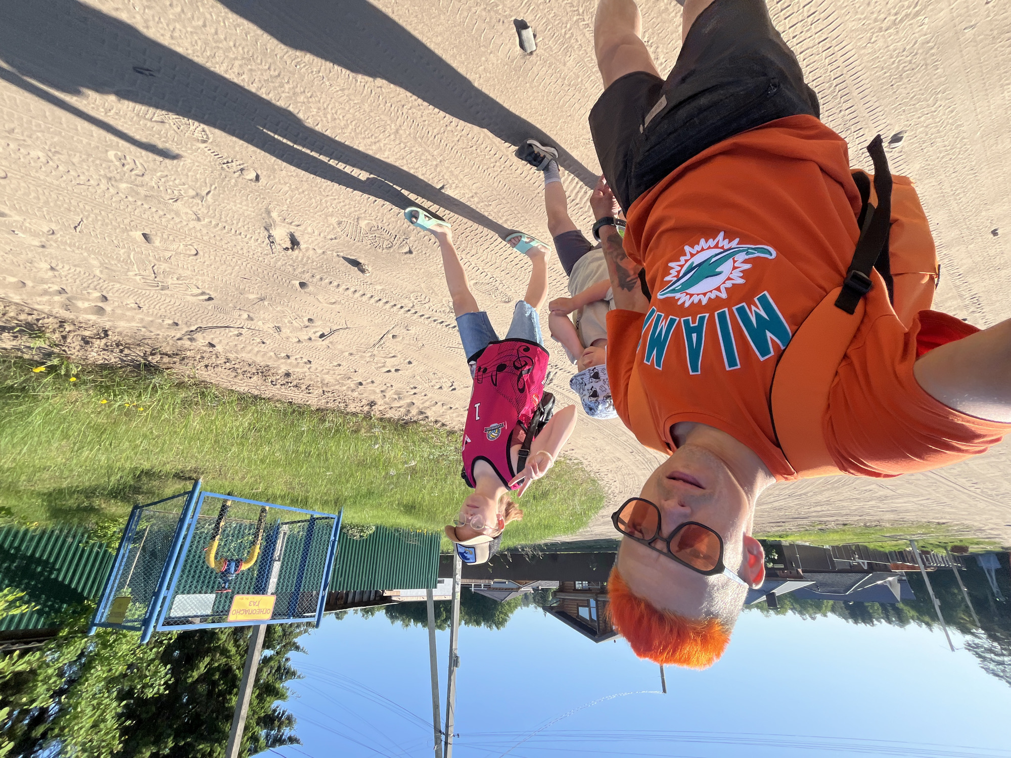 A man with orange hair and sunglasses wears an orange "Miami" shirt while holding a young child, along with a teenager in a pink jersey, on a sandy path near a green corrugated fence and houses. The location is Chaikovsky, Russia.