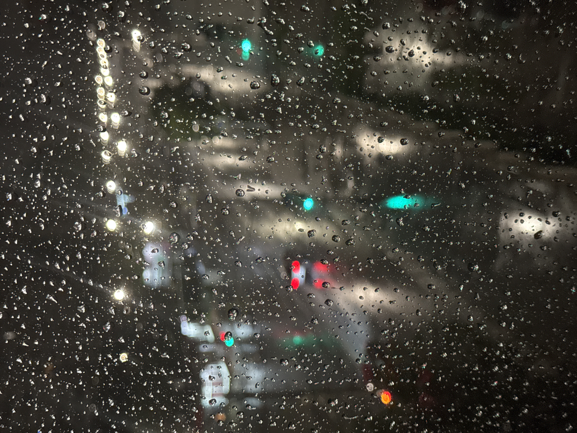 The image shows a rain-streaked window at night, with blurred lights visible through the droplets. The light sources suggest a street scene with traffic signals (green and red), car headlights, and streetlights. The overall impression is of a rainy night in an urban environment. The location, Saratov, Russia, cannot be confirmed. No people, subjects, objects, actions or interactions are visible.