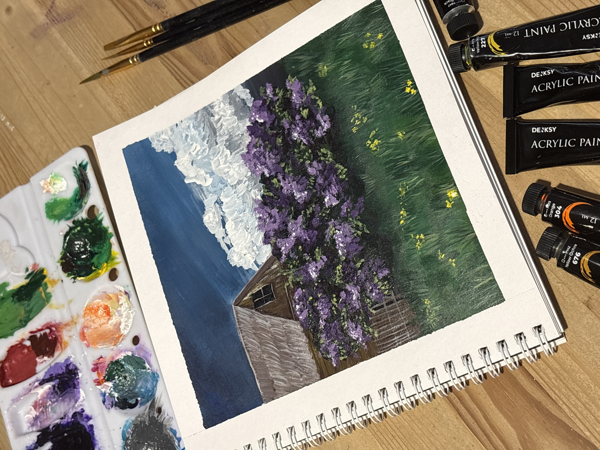This overhead shot, taken in Perm, Russia, captures an active acrylic painting setup on a wooden surface. The central focus is a vibrant, impressionistic painting in a spiral-bound sketchbook.
The artwork depicts a serene rural landscape. A rustic wooden building, possibly a barn or house, with brown siding and a small, dark window, is partially covered by a dense, flowering purple vine, likely wisteria or lilac. Above, a deep blue sky is rendered with thick, textured white clouds, showcasing distinct brushstrokes. To the right, a lush green field is interspersed with small yellow flowers.
Surrounding the painting are various art supplies. To the left, a white plastic artist's palette holds an array of mixed acrylic paints, including shades of green, orange, red, purple, blue, black, and white, indicating an ongoing creative process. Above the palette, three fine-tipped paintbrushes with black handles and gold ferrules rest on the wooden table. On the right, several black tubes of "DEJKSY ACRYLIC PAINT" are visible. Text on the tubes identifies some colors as "Yellow Mo 227", "Yellow Ochre 676", and "Orange 304", along with pigment codes like "+++PY42" and "+++P013" and the volume "12 ML". The overall scene evokes a sense of artistic concentration and creativity.