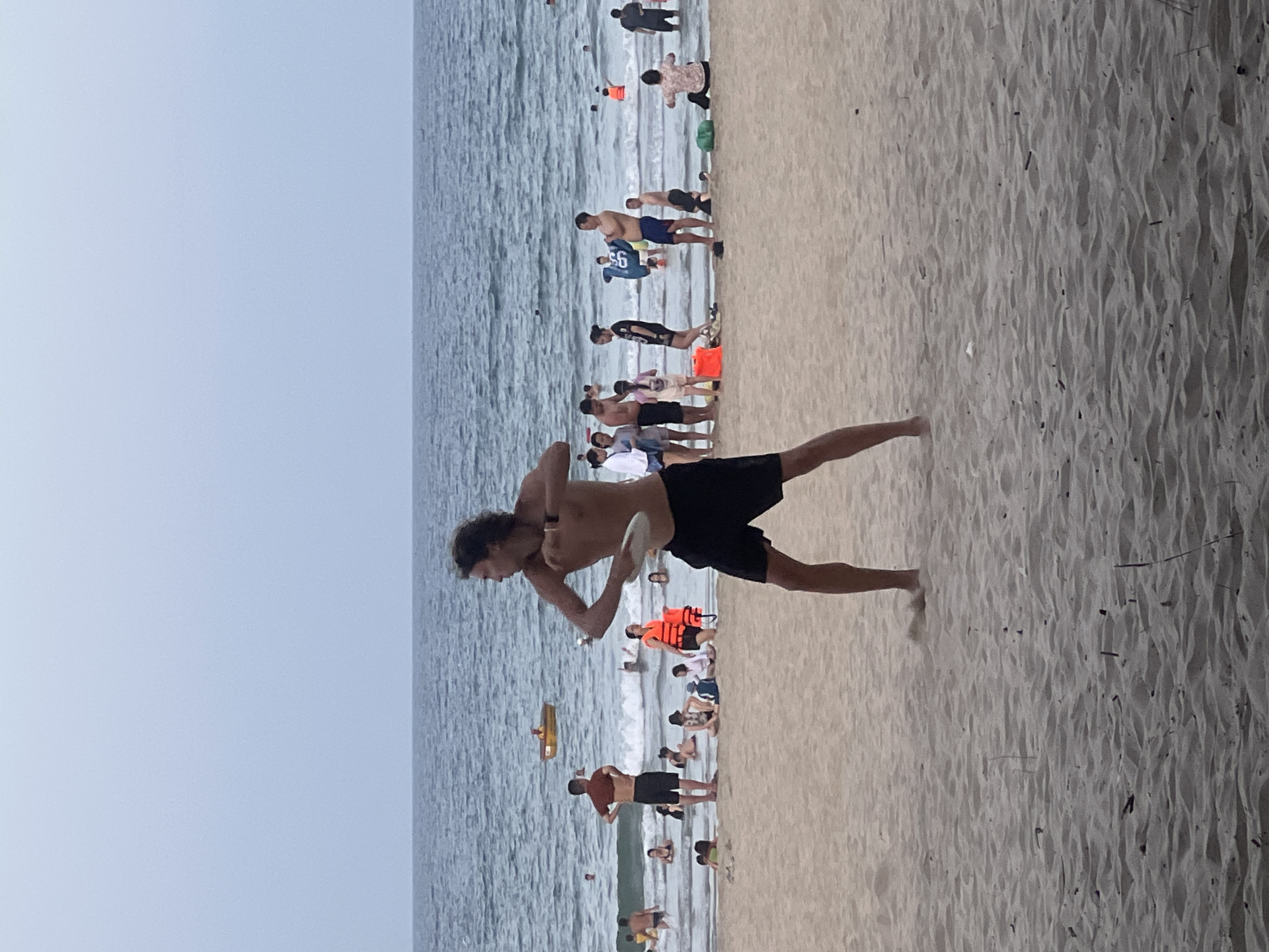 The image shows a crowded beach scene at Đà Nẵng, Vietnam, on an overcast day. The foreground is dominated by a man with dark, curly hair, wearing black shorts, who is actively engaged in a game of paddleball, holding a white paddle in his right hand. His body is angled towards the left of the frame, in mid-action.
Further back, on the sandy beach and in the shallow water, numerous other people are visible. Some are wading in the sea, while others are sitting or standing on the sand. There are groups of people, including families and individuals.
In the background, the ocean stretches to the horizon under a pale sky. Small waves are breaking near the shore, and a few boats are visible in the water. One prominent boat, a yellow, shallow-draft vessel, is near the center of the frame with at least one person in it. The overall atmosphere is one of leisure and recreation on a busy beach.