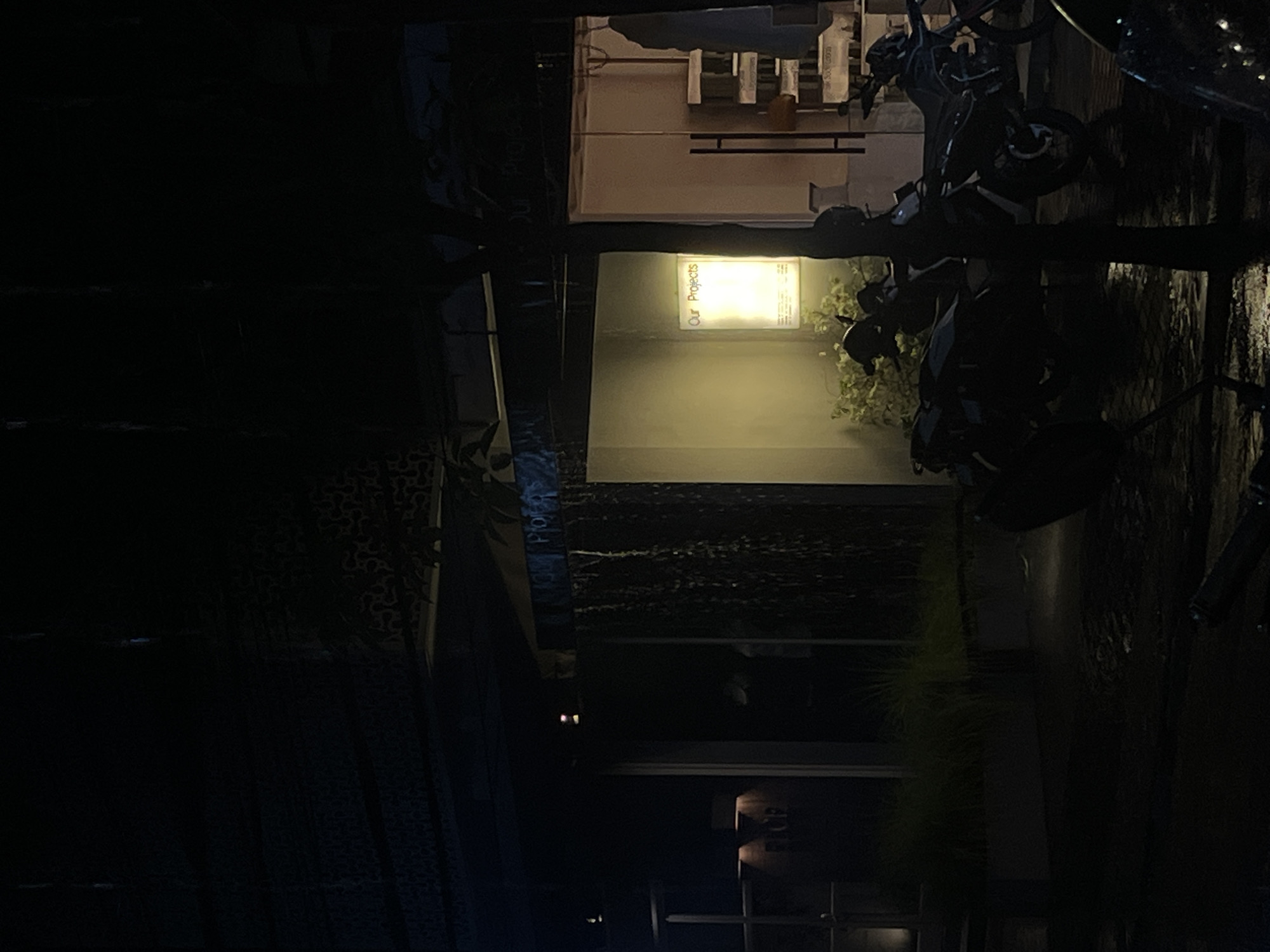 The image depicts a nighttime scene in an urban or semi-urban environment, likely in Mengwi, Indonesia, as indicated by the location context. The central focus is a brightly illuminated rectangular sign on a building, displaying the text "Our Projects" vertically in white lettering against a warm yellow light. Green foliage or plants are visible around the base and to the right of the sign.
To the right of the illuminated sign, a dark motorcycle or scooter is partially visible, parked on what appears to be wet pavement or ground. Above the sign and behind the vehicle, a glimpse into a building's interior reveals what looks like shelving or architectural details under artificial light. The left side of the frame and much of the foreground and background are cloaked in deep shadow, hinting at other dark building facades or structures. No people are visible, and there is no specific activity taking place; the scene is static and quiet, illuminated primarily by the sign's light. The overall atmosphere is dark, with the artificial light creating a focal point against the night.