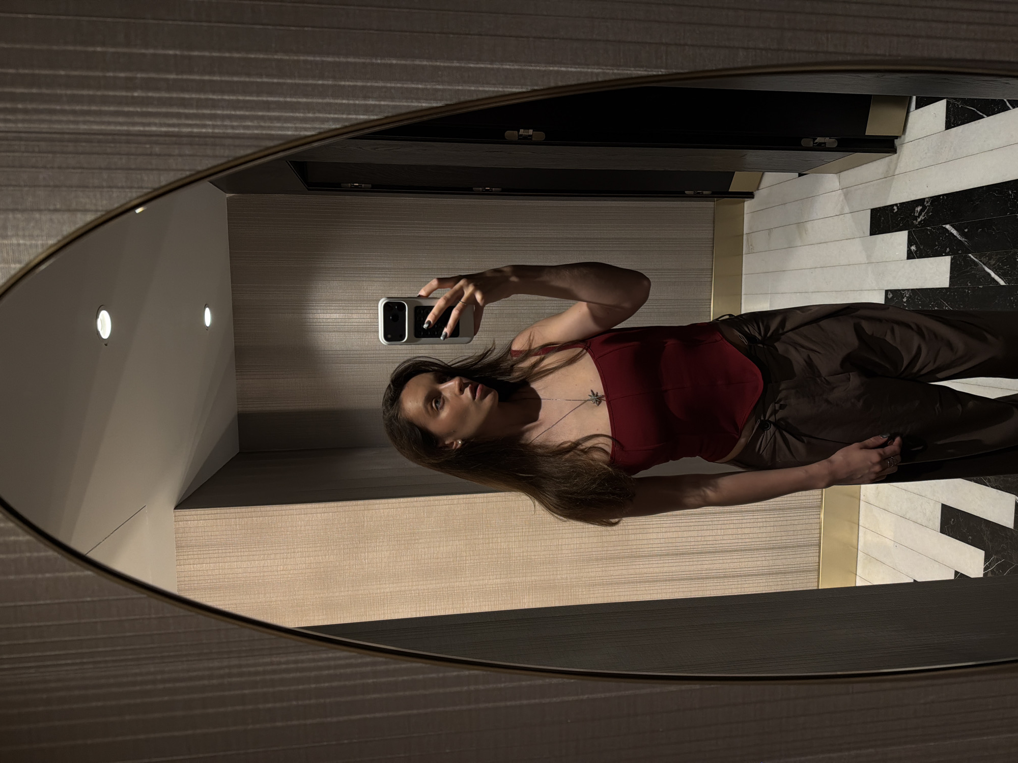 A woman is taking a selfie in a mirror in what appears to be a bathroom. The mirror is oval-shaped and has a gold frame. The woman is wearing a maroon top and brown pants. She is holding a phone in her right hand. The background includes a light-colored wall with horizontal lines and a black-and-white patterned floor. The scene is dimly lit. This picture was taken in Budva, Montenegro.