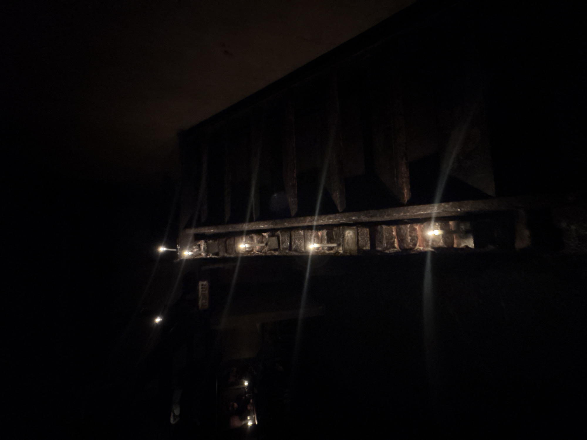 The image depicts a very dark scene, likely an interior at night or in extremely low light conditions. A prominent horizontal structure, possibly a wooden or metal beam or ledge, spans across the middle ground. Along this beam, several small, bright white lights are affixed, creating distinct lens flares that emanate from each light source, emphasizing the low-light environment.
Above the illuminated beam, dark vertical elements, resembling balusters or slats, are visible, suggesting part of a railing or a decorative partition. The material appears rugged or aged. The area below the beam is almost entirely enveloped in deep shadows, with only faint, indiscernible shapes hinted at within the darkness. A solitary light source is also present on the far left, detached from the main cluster on the beam.
No people are visible, and no specific activity is taking place; the image captures a static arrangement of lights and structure. The time of day is clearly night or a similarly dark indoor setting. The atmosphere evoked is one of intimacy, mystery, or coziness, primarily due to the contrast between the intense darkness and the small, bright points of light. While a small, rectangular object can be seen hanging below the beam on the left, any text on it is illegible due to blurriness and shadow. Given the location context of Sofia, Bulgaria, this could be an architectural detail in a restaurant, bar, or a building interior.