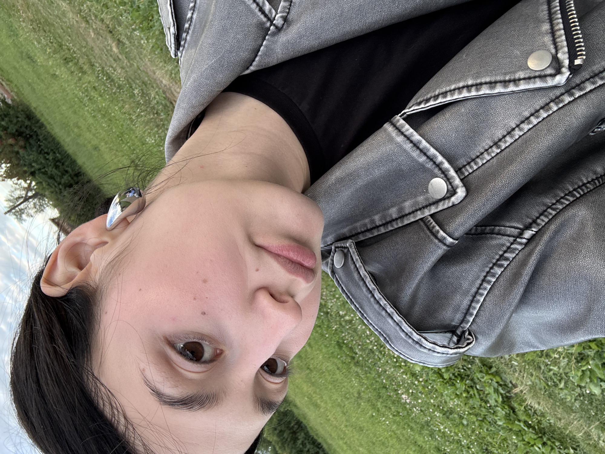 The image features a close-up selfie of a young woman outdoors, with her head tilted and facing the camera. She has dark hair, brown eyes, and light skin with some freckles visible. She is wearing a black t-shirt and a grey faux leather jacket with prominent stitching and silver snap buttons. A silver, pointed earring is visible on her left ear. The background is blurred, showing a green grassy field with some sparse white flowers, suggesting an outdoor setting in daylight. The lighting indicates it could be late afternoon or early evening, with soft, natural light. The overall mood appears casual and relaxed. There is no visible text in the image.