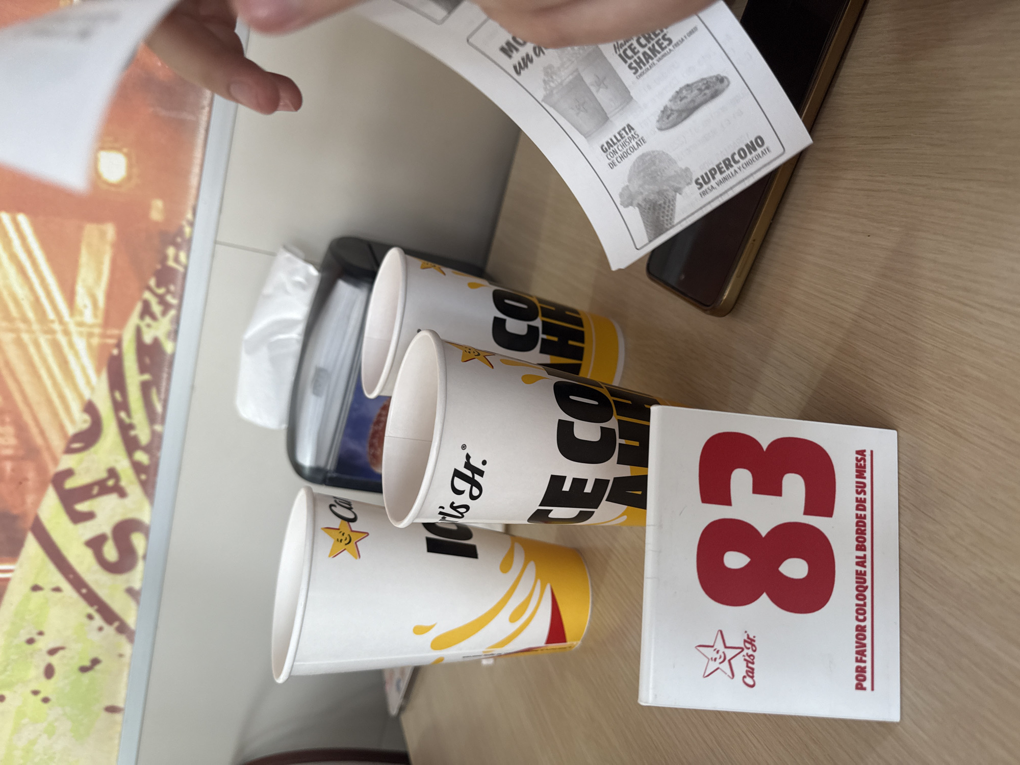 The image shows a table at a Carl's Jr. restaurant. There are three Carl's Jr. cups, a number stand showing the number 83, a menu, a phone, and a tissue box. The menu lists ice cream shakes and a "Supercono". The number stand has the Carl's Jr. logo and the phrase "POR FAVOR COLOQUE AL BORDE DE SU MESA". Based on the OCR text "CE COAH", the cups likely contain Carl's Jr. Cola. A hand is visible holding the menu.