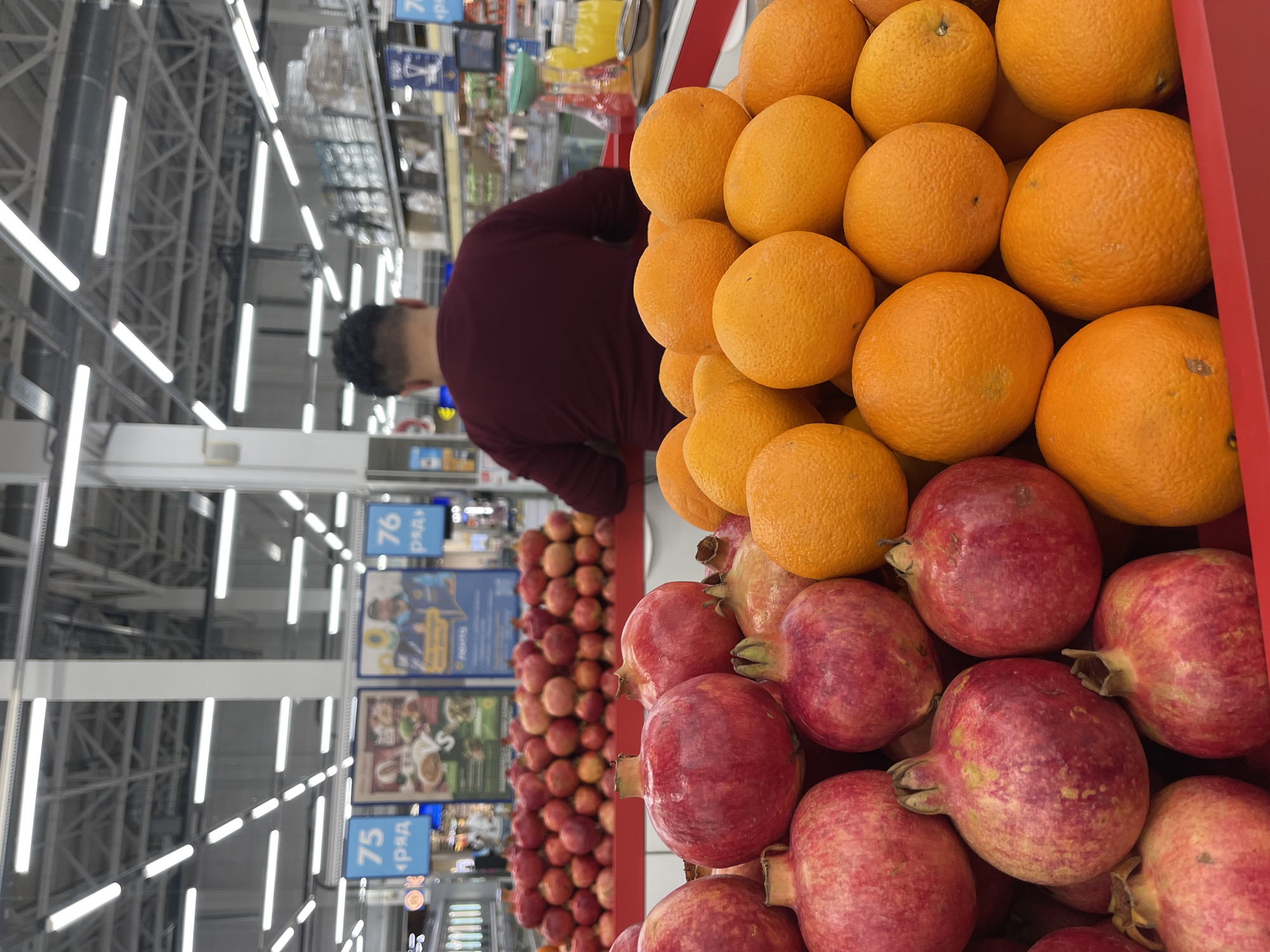 The image shows a produce display in a supermarket in Shcherbinka, Russia. In the foreground, there are oranges stacked above pomegranates in a red bin. To the left, there is a person with short dark hair, facing away from the camera, wearing a maroon shirt. The aisles of the supermarket stretch into the background, with overhead lighting and numbered signs indicating rows. Apples can be seen in the left background.