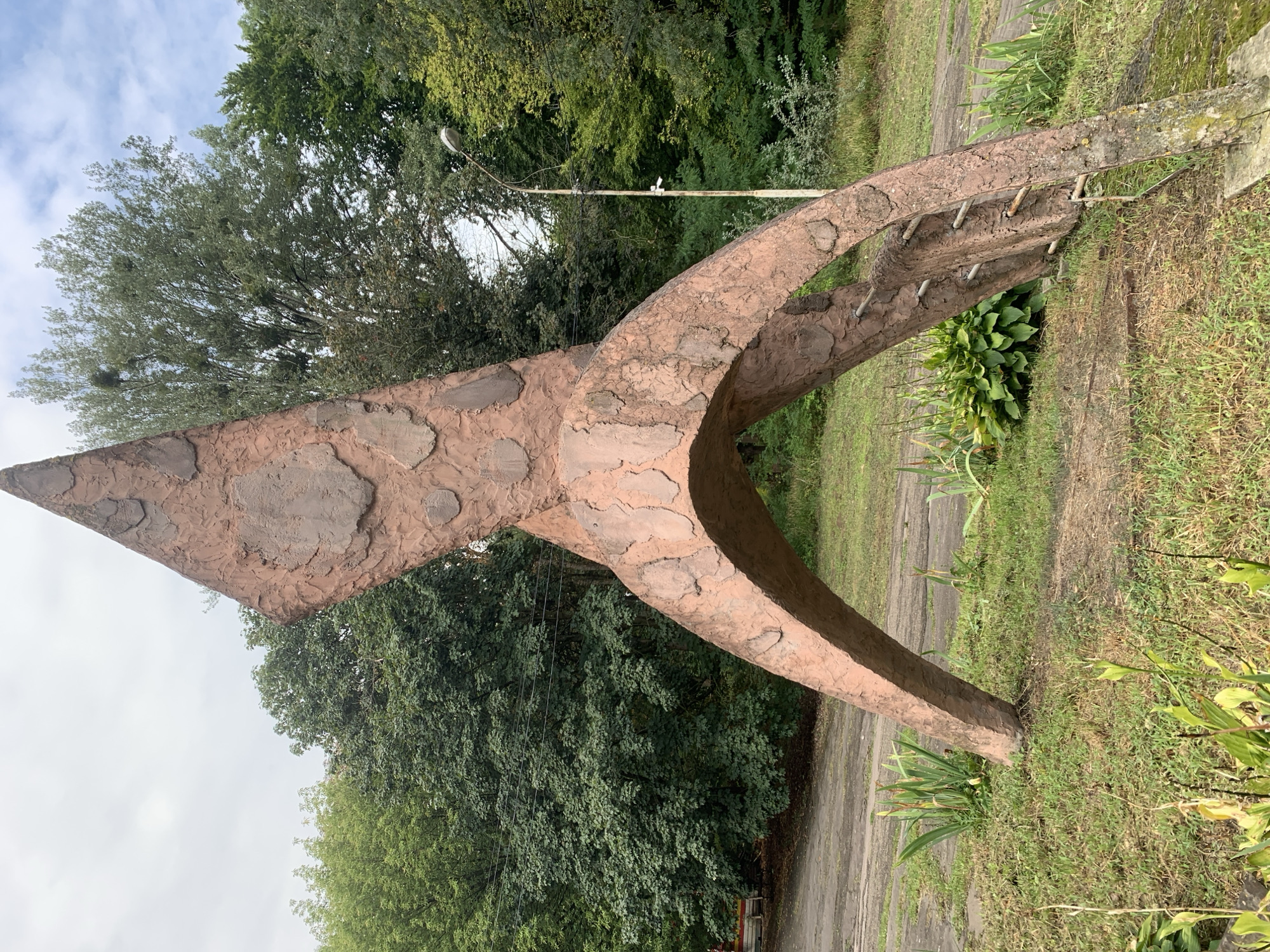 A large, abstract concrete sculpture, with a rough, textured surface resembling bark or stone, stands in a grassy area. The sculpture is predominantly reddish-brown and has a dynamic, upward-sweeping form, with an arching base. One side of the sculpture appears to have metal rungs embedded in it, suggesting it might have been intended as a climbing structure or has weathered to show internal reinforcement.
The sculpture is situated outdoors, with lush green trees forming a dense backdrop. The ground around the sculpture is covered in grass and some low-lying plants. A dirt or gravel path is visible behind the sculpture. In the background, a street lamp on a tall, thin pole stands among the trees, and power lines are strung between trees. The sky is overcast with diffused light.
The style of the sculpture suggests it could be from the Soviet era, a common period for such public art in Eastern Europe. While the exact location is not specified in the image itself, this type of monument is reminiscent of many found in parks or public spaces in cities across Ukraine, including Lviv. Without specific markings or contextual information within the image, pinpointing Lviv with certainty is not possible, but it is plausible given the aesthetic. There are no people visible in the image.