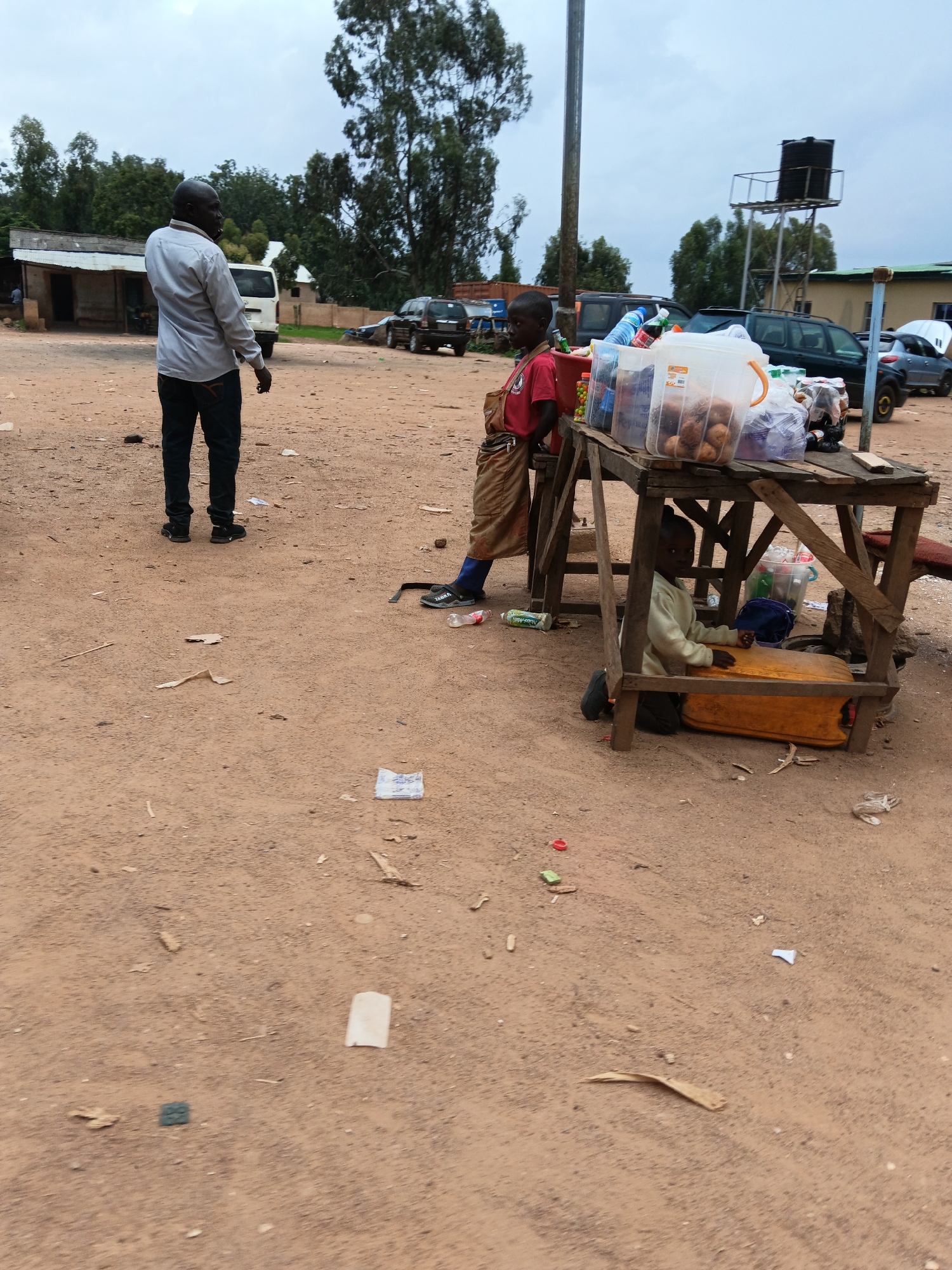 An outdoor scene in an urban environment, likely a marketplace or roadside stall, on an overcast day. A man stands on the left, facing away from the camera, wearing a light-colored collared shirt and dark pants. He appears to be talking on a mobile phone. To the right, a wooden market stall displays various items, including plastic containers filled with food items like bread rolls, bottled drinks, and other goods. A young boy, dressed in a red shirt and brown shorts, stands behind the stall, leaning against it. Underneath the stall, another young boy is seated on the ground, wearing a light-colored long-sleeved shirt and dark pants, looking towards the camera. The ground is dirt and there are various discarded items, such as plastic bottles and paper scraps, scattered around the scene. In the background, several vehicles are parked, including a white van, a dark SUV, and a gray car. Buildings and trees are visible in the distance, along with a water tower.