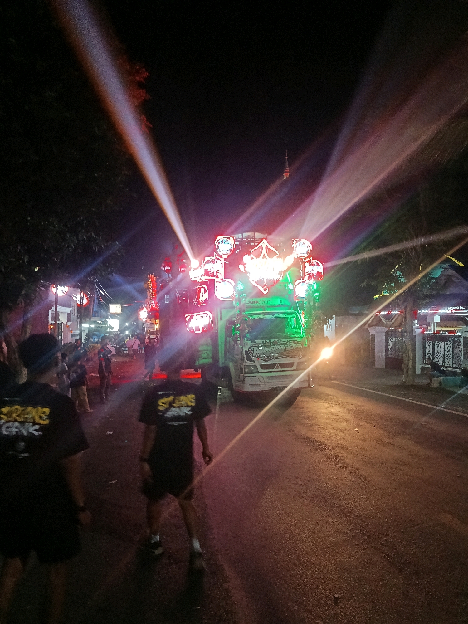 In Pasirian, Indonesia, a vibrant night scene unfolds on a street illuminated by a dazzling array of lights emanating from a truck adorned with a large sound system and multiple spotlights. The truck, a central element, is packed with speakers and lights, casting beams of white, red, and green light into the dark sky, creating dramatic streaks and flares. The light show is further enhanced by smoke or haze, which catches the light and adds to the atmospheric effect.
Several individuals are present, with two men in the foreground wearing black t-shirts with yellow lettering. They appear to be walking away from the camera, towards the illuminated truck. Other people are scattered throughout the scene, some gathered near the truck and others further down the street, suggesting a gathering or event. The street itself is asphalt, and the surroundings suggest a residential or semi-urban area, with buildings visible on either side and trees lining the road. The overall impression is one of a lively and festive atmosphere, likely a cultural event or celebration.