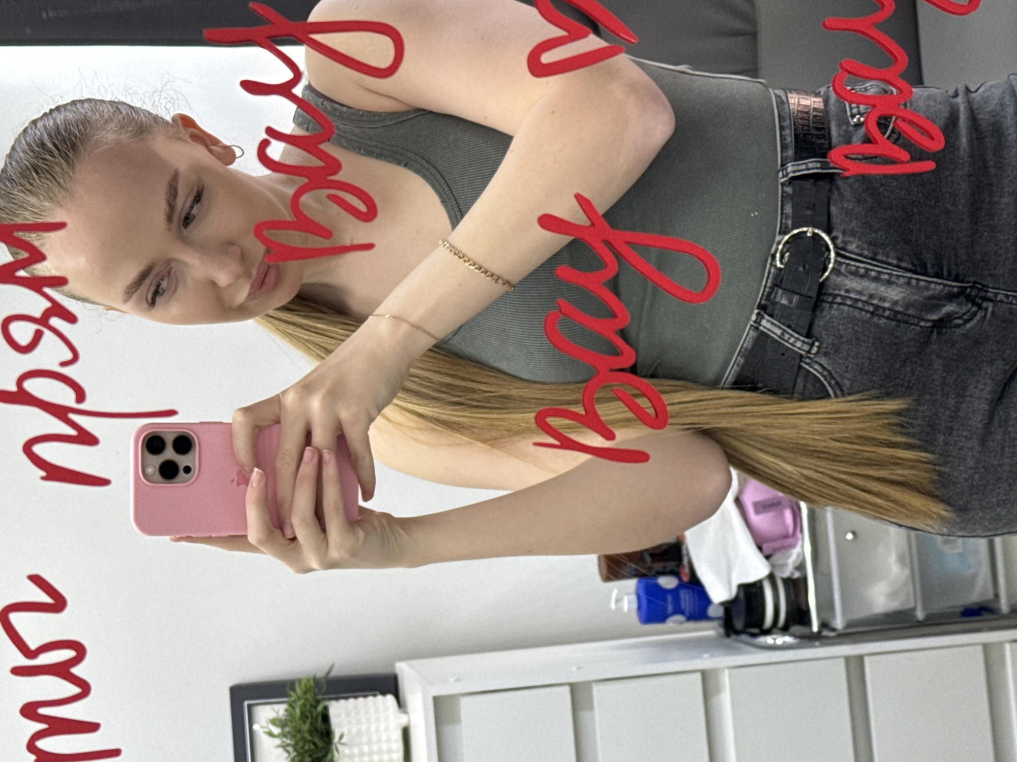 A female individual is captured in a mirror selfie within an indoor setting in the city of Petropavlovsk-Kamchatsky, Russia. The subject, appearing to be an adult with fair skin and light-colored hair pulled into a low ponytail, is wearing a dark grey ribbed tank top and dark grey washed denim jeans secured by a black belt with a silver D-shaped buckle. A thin gold-colored chain bracelet is visible on her right wrist, and a small hoop earring adorns her left ear. She holds a pink-cased smartphone with a multi-lens camera module in both hands, directing her gaze towards the screen with a subtle smile. Red, stylized script text is affixed to the mirror's surface, partially overlaying her reflection; fragments read "ты прям" (ty pryam - "you're really") at the top and multiple instances of "Вау" (Vau - "Wow") are visible in the mid-ground and lower right. The reflection reveals a white multi-drawer storage unit on the left, topped with a black-framed object and a potted green plant in a white textured container. Behind the subject's right arm, a blue pump bottle, a dark brown container, and a white cloth are visible on or near a small, metal-framed storage cart with drawers, which also holds a pink object. The room's walls appear to be white.