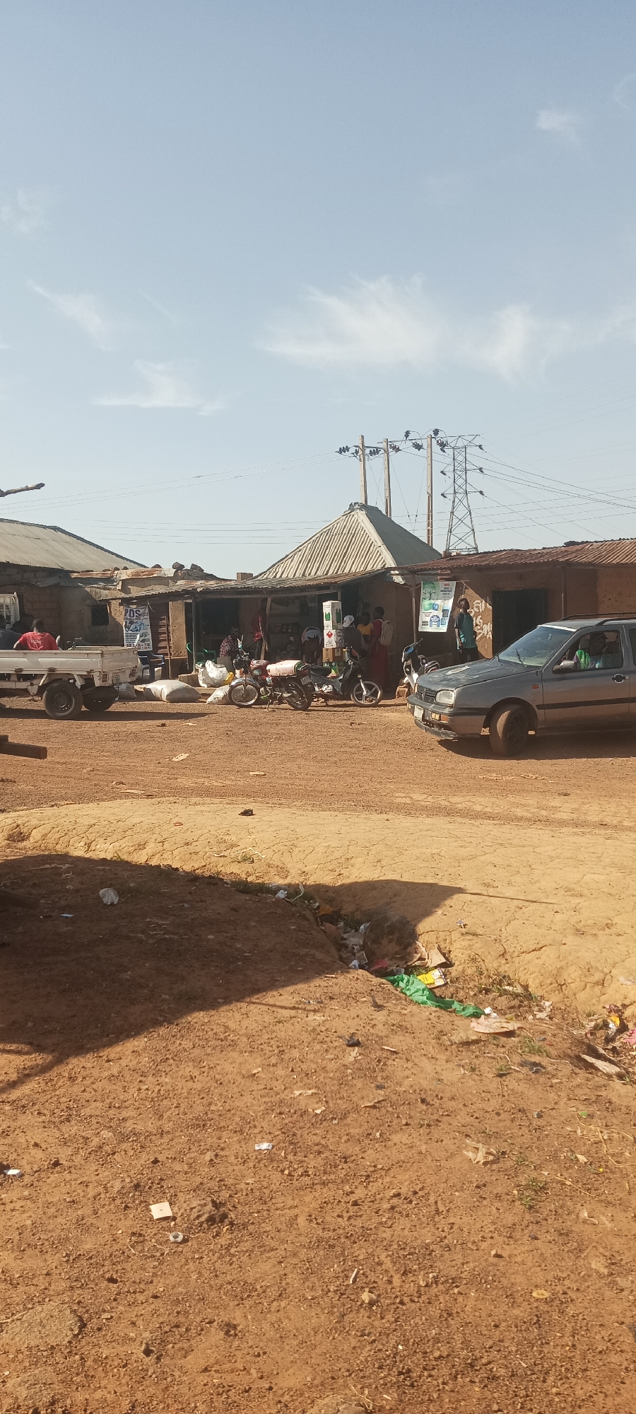 The image shows a street scene in an unspecified location of Unknown, Unknown. A small, dirt road dominates the foreground with scattered litter and debris. A silver car is parked on the right side of the road, and a white pickup truck is on the left. Several motorcycles are parked in front of a small shop or market. A group of people appear to be gathered at the shop. Power lines are visible in the background. The sky is clear. Buildings along the road appear to be constructed with corrugated metal roofs and earthen walls.
