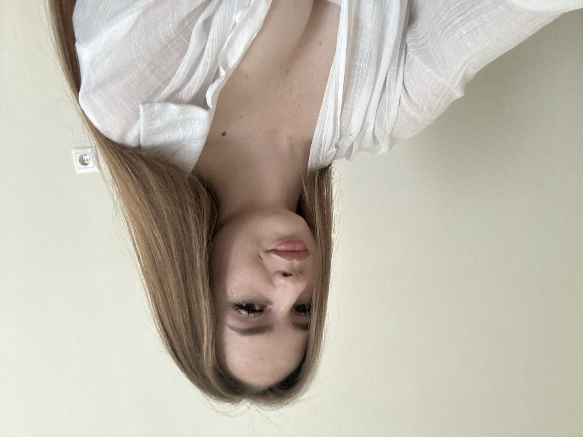 A young woman with long, light brown hair is pictured indoors, looking towards the camera. She is wearing a white, open-collared shirt, revealing her décolletage. The background is a plain, light-colored wall with a white electrical outlet visible on the right side. The lighting is soft and natural, illuminating the woman's face and the wall behind her.