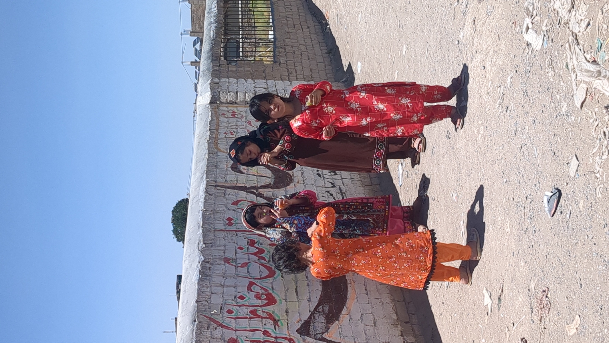 The image displays four young girls standing on a sunlit, unpaved street in what appears to be a residential area of Quetta City Tehsil, Pakistan. They are positioned in a line, with a low wall behind them and a clear, bright blue sky above.
The girls are dressed in traditional South Asian attire, consisting of long-sleeved, patterned dresses and loose pants. From right to left:
The first girl wears a red dress with white floral patterns and dark sandals. She looks towards the camera with a neutral expression, holding a small object in her hand.
The second girl is in a dark brown dress with embroidered details and a dark head covering, which partially obscures her face as she looks downwards.
The third girl wears a vibrantly patterned, multi-colored dress and a pink headscarf, making a peace sign towards the camera.
The fourth girl, dressed in an orange floral-patterned dress and orange pants, stands slightly turned away, looking towards the group.
The ground is a light sandy or dirt surface, strewn with scattered litter, including plastic pieces and other debris. The strong, distinct shadows cast by the girls indicate direct sunlight, suggesting it is mid-morning or afternoon on a clear day. The wall behind them is made of concrete or brick, partially covered in white plaster, with visible red and black script, likely in Urdu or Farsi, applied as graffiti or artwork. In the distance, above the wall, a green tree is visible to the left, and some building structures are faintly seen to the right. The scene captures a casual, everyday moment in a local community setting.