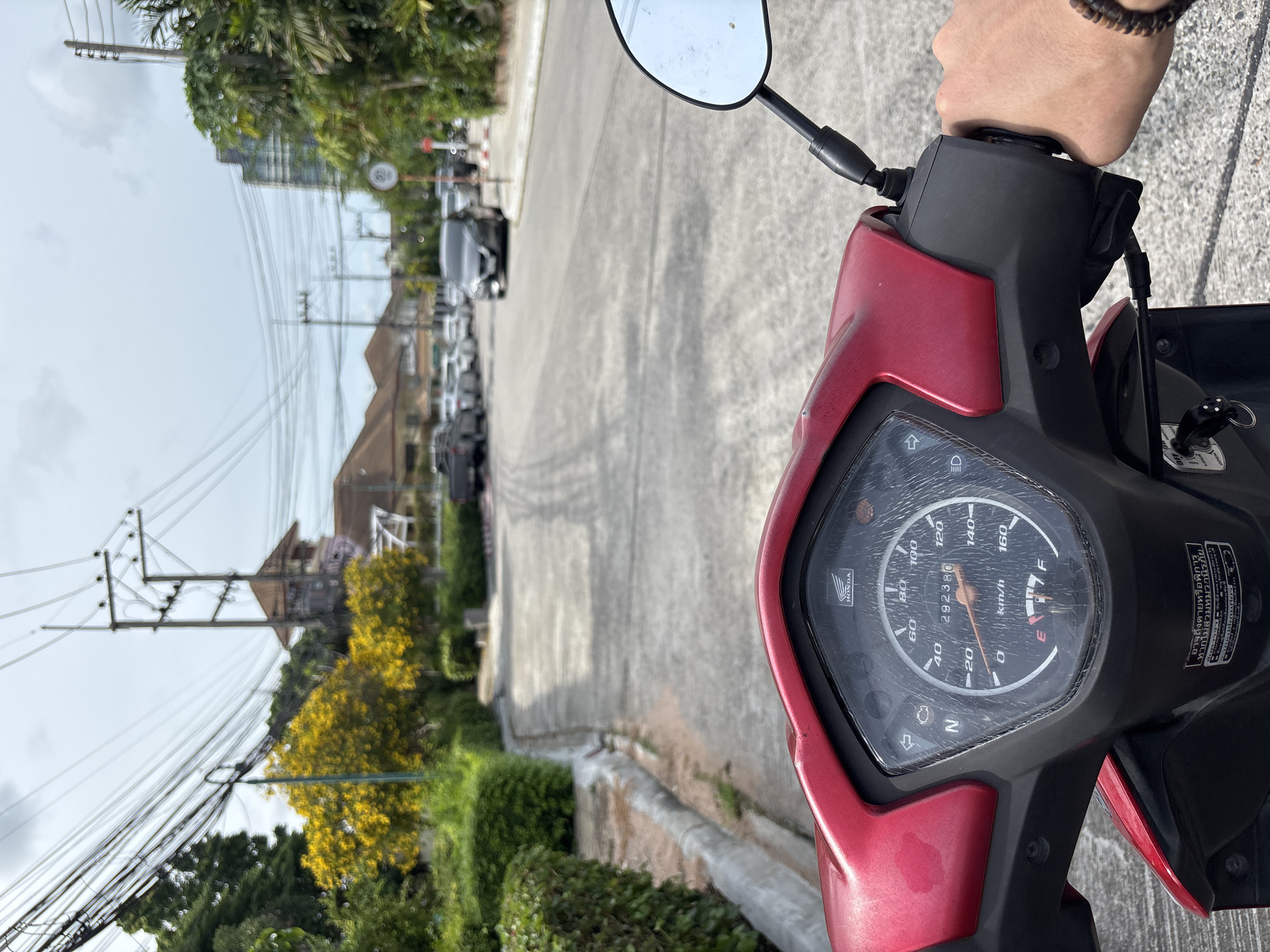The image, taken from the perspective of someone riding a red Honda motorcycle, shows a street scene in Pattaya City, Thailand. The motorcycle's dashboard is in the foreground, displaying a speedometer with a needle pointing to approximately 20 km/h and an odometer reading 292380. The rider's right hand is visible on the handlebar, wearing a bracelet.
The street is paved with concrete, with a slight curve to the left. A concrete curb runs along the left side of the road, bordered by dense green bushes. In the background, a line of parked cars is visible on the right side of the street, and a speed limit sign (20) is posted on a pole. Further down the road, more buildings and trees can be seen, including a tall building and houses with tiled roofs. Overhead, a complex network of power lines crisscrosses the sky, supported by utility poles. Yellow flowering trees are prominent on the left side of the road, adding a splash of color to the scene. The sky is overcast.