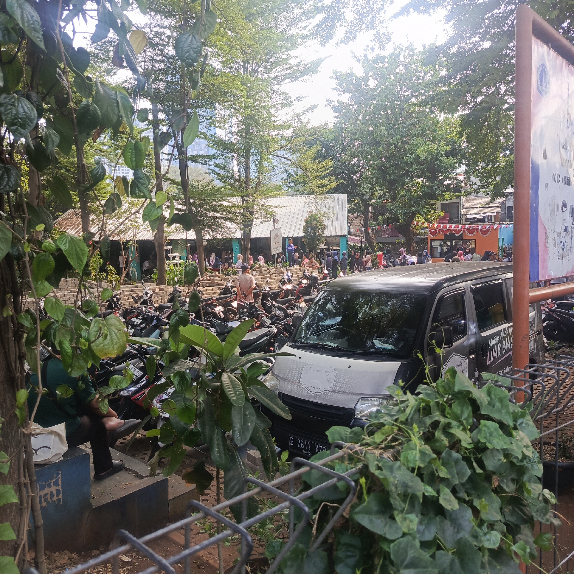 The image depicts an outdoor scene, likely in a market or public gathering area, with a clear presence of parked motorcycles and a vehicle. In the foreground, lush green foliage partially obstructs the view of a dark-colored van. The van has text on its side, including "LUXURIOUS" and license plate "B 2811 KZD." Behind the van, numerous motorcycles are parked closely together.
In the middle ground, a structure with a corrugated metal roof is visible, possibly a market stall or food court, with people seated at tables. A pile of bricks is arranged in front of this structure. Several individuals, many wearing headscarves, are seen sitting or standing in and around this area.
The background features tall trees, some with buildings or structures peeking through their branches, suggesting an urban environment. Sunlight filters through the leaves, creating a bright and slightly overcast sky. A large signpost with some text and graphics is visible on the right side of the image. The overall impression is a busy, casual outdoor space in what appears to be South Jakarta, Indonesia, based on the license plate format.