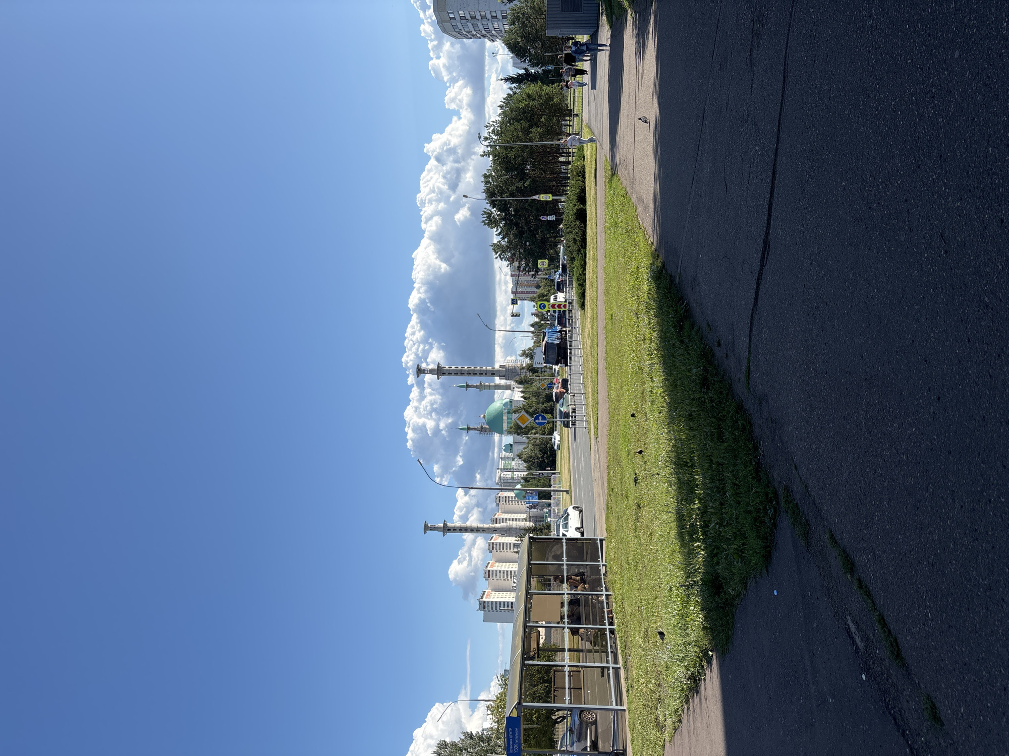 This is a medium-angle, full-shot photograph of a sunny day in Naberezhnye Chelny, Russia. In the foreground, a paved walkway leads away from the viewer, bordered by a grassy area. To the left, a bus stop shelter with a glass enclosure stands, and a few people are seated inside. Beyond the bus stop, a street is visible with cars and a bus. In the middle ground, there are trees and more buildings, including a prominent mosque with a green dome and tall minarets. The sky is a bright blue with scattered white clouds. The scene depicts a typical urban environment with infrastructure for public transportation and religious buildings.