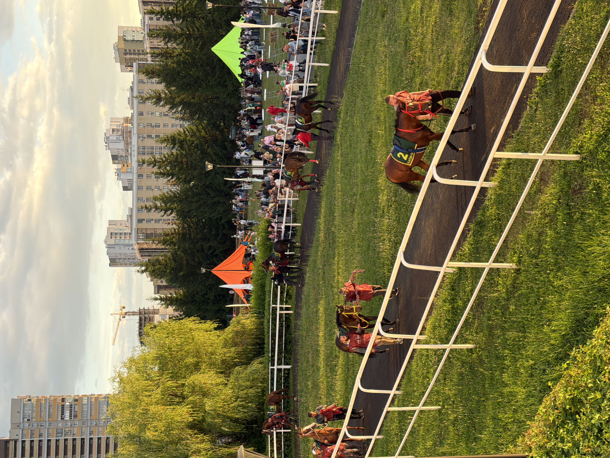 In an outdoor urban setting at golden hour, several horses are being led along a dirt track, bordered by white railings and a grassy slope. People in red shirts, likely handlers or jockeys, are walking with the horses. One horse in the foreground has the number "2" and the word "КАЗАНЬ" visible on its saddle pad. In the background, a crowd of spectators gathers behind a railing, with several large tents, including an orange one and a green one, set up in the event area. Behind the event, a line of modern apartment buildings and a construction crane are visible against a cloudy sky. The lighting suggests it is late afternoon or early evening.