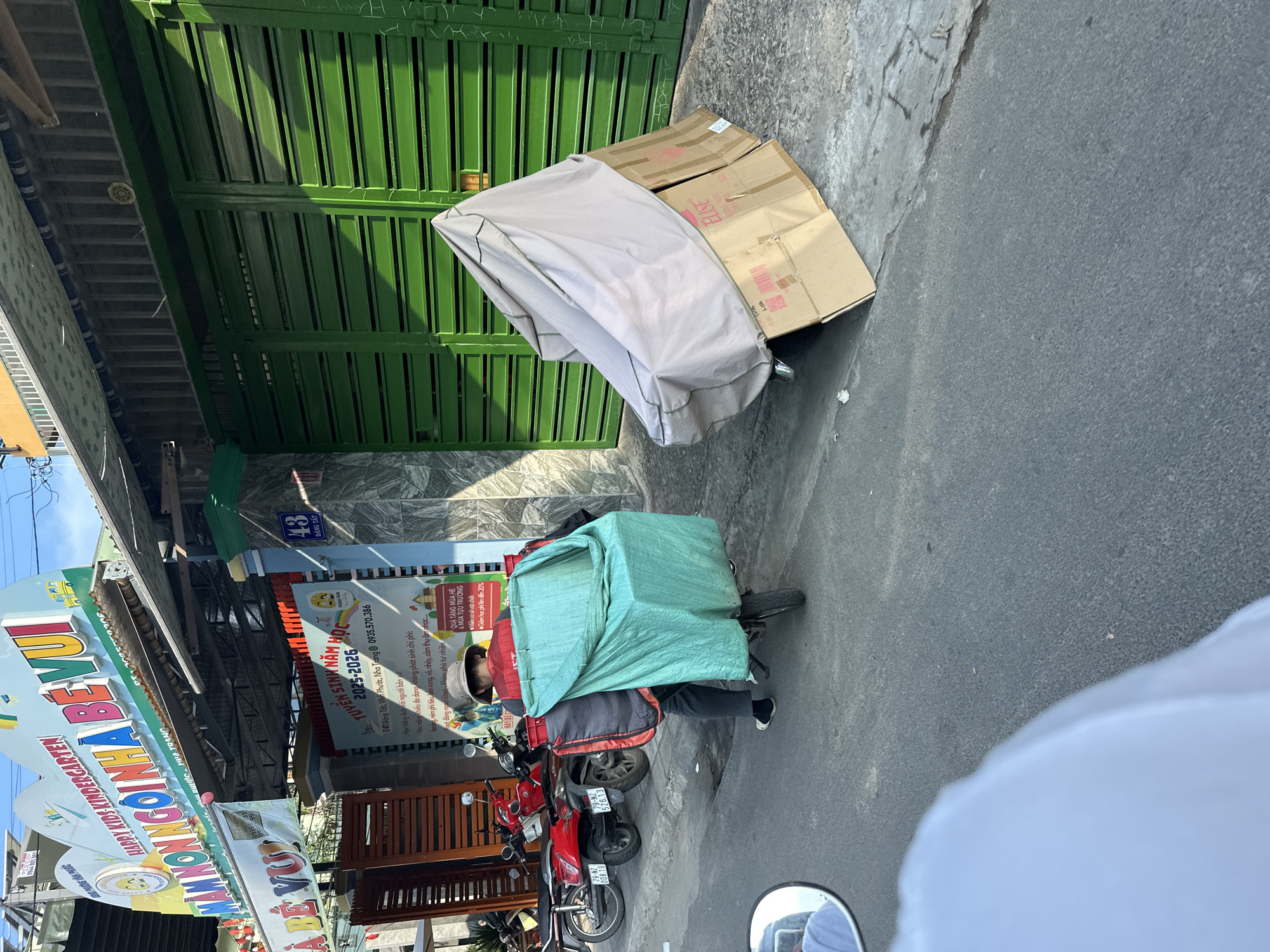 The image captures a street scene in Bac Nha Trang Ward, Vietnam. A delivery person on a motorcycle, covered by a green tarp, is parked near a green metal gate. To the gate's right is a large package and cardboard box. A kindergarten's sign, "HAPPY KIDS KINDERGARTEN," is visible, along with another sign with a phone number. Other parked red motorcycles are on the side. The image appears to be taken from another moving vehicle, focusing on the delivery person and the immediate surroundings.