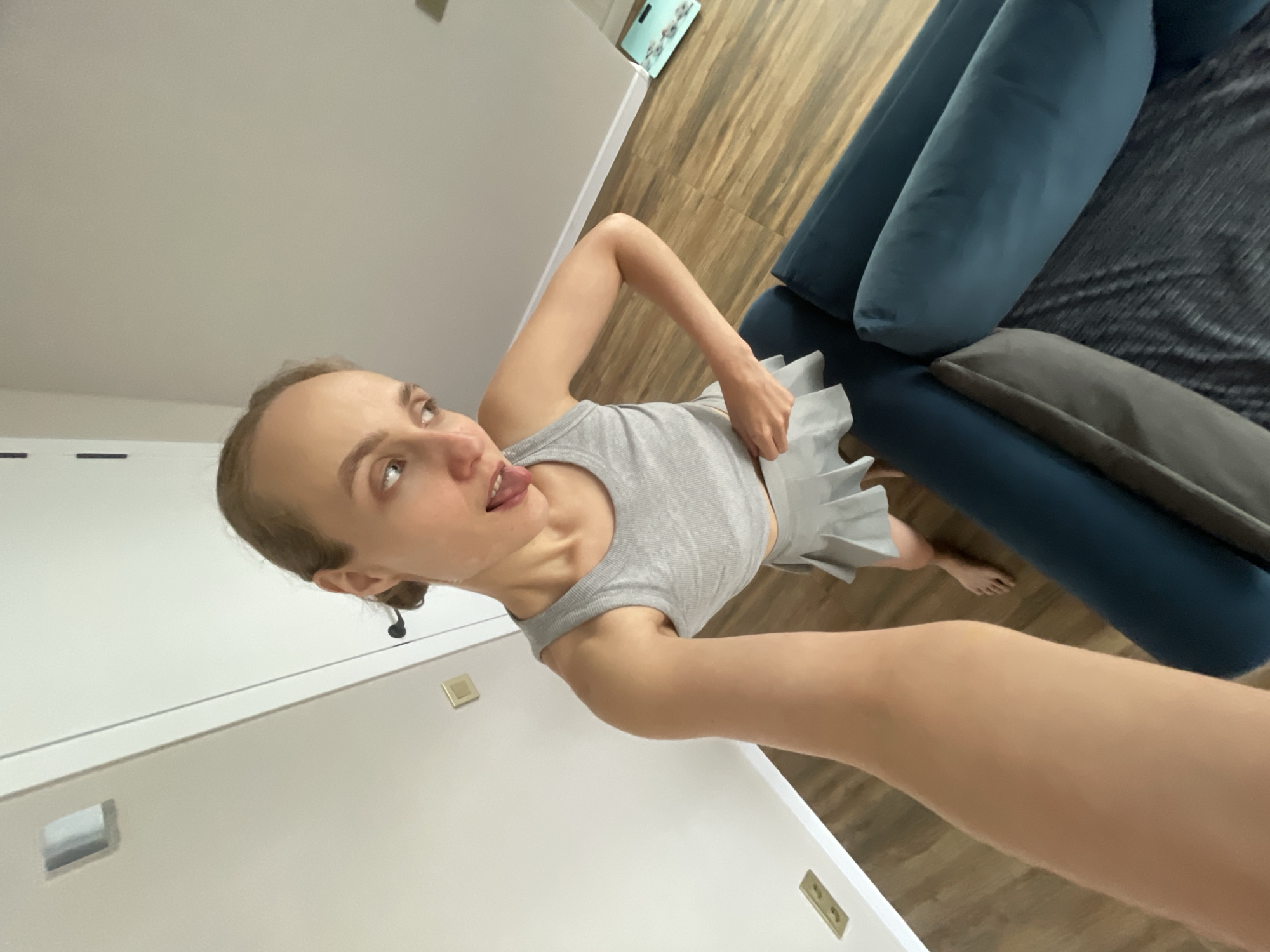 The media file shows a young woman indoors, possibly in a living room. She is the sole subject, wearing a grey tank top and a grey pleated skirt. She is looking upwards and to the side with her tongue sticking out playfully. Her hair is pulled back into a bun, and she is holding a white piece of fabric or paper. The background consists of a wooden floor and a teal sofa with several cushions. There are also some wall features that appear to be light switches or electrical outlets. The lighting suggests it could be daytime. The overall mood seems lighthearted and candid. There is no visible text in the image.