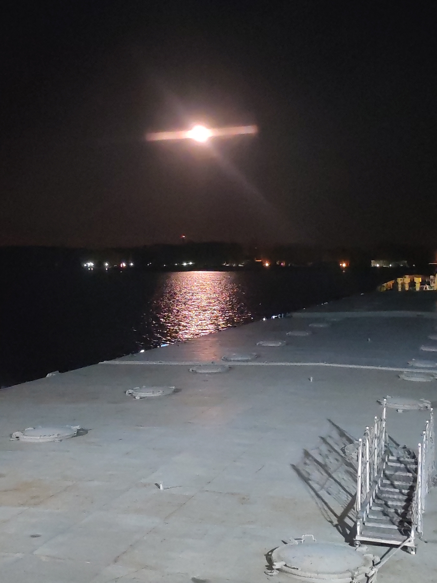 The image depicts a nighttime scene, possibly taken from the deck of a ship or structure near water. The sky is dark with a bright light source, likely the moon, prominently visible and reflected on the water's surface. The water shows a shimmering, reddish reflection of the light source.
The foreground features a gray surface with several round, metal hatches and a set of metal stairs or scaffolding casting long shadows. In the distance across the water, there are faint lights indicating a shoreline or another structure.
The location is labeled Unknown, Unknown.