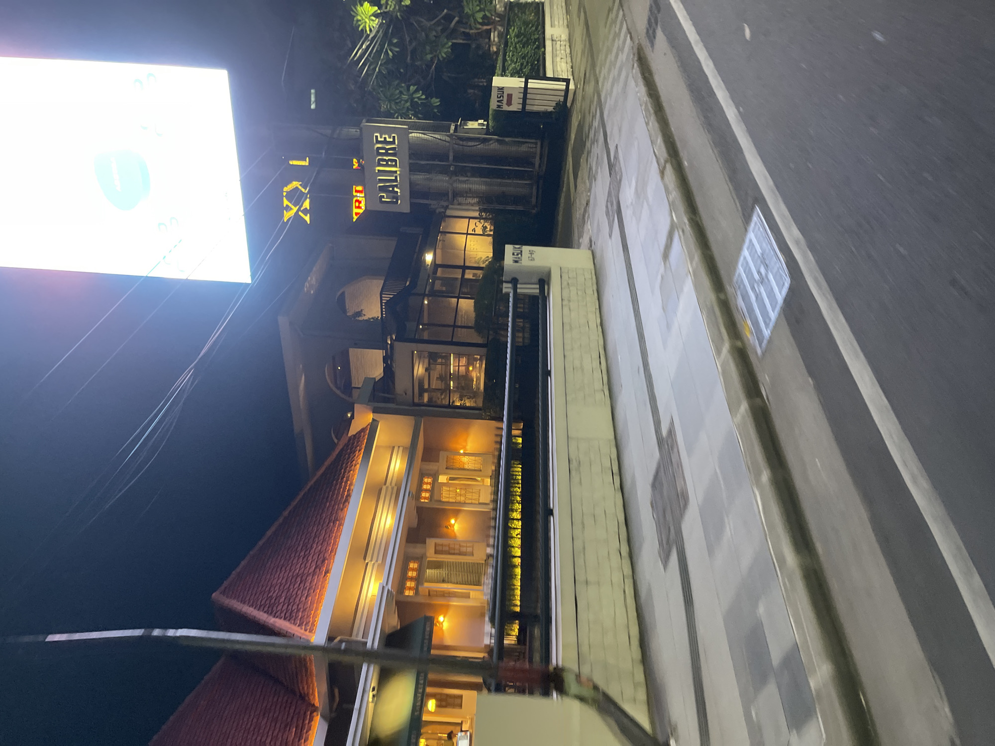 The image shows a street view at night in Surabaya, Indonesia. On the right side of the image, a lit sign reads "CALIBRE." The building behind it has large windows and a dark interior. To the left of the "CALIBRE" sign, a smaller sign with an arrow indicates "MASUK" (Indonesian for "Enter"). Further left, a building with a red-tiled roof is illuminated from below with warm yellow lights. The street in front of the buildings has a sidewalk with square grates and a curb. The street itself is paved and appears wet, reflecting the streetlights. The overall impression is of an entertainment or dining establishment in a bustling urban area.