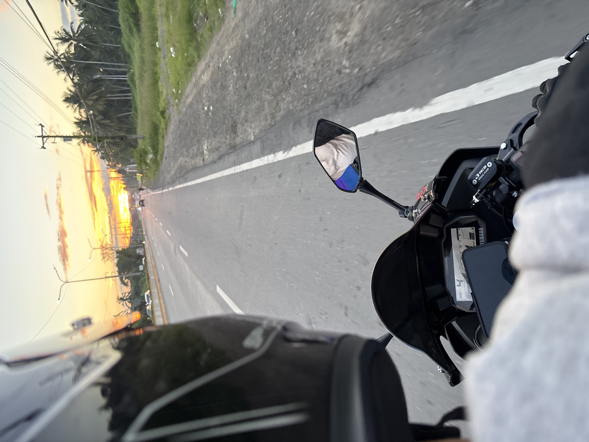 A first-person perspective shot from a motorcycle traveling on a road during sunset. The rider is wearing a black helmet with white stripes and a gray jacket. In the foreground, the motorcycle's dashboard and handlebars are visible, with a phone mounted on the handlebars. The road stretches ahead, with a white line marking the lane. To the right of the road is a grassy area with scattered litter and a row of palm trees. In the distance, the sky is ablaze with the warm colors of the setting sun, casting a glow over the scene. A few vehicles are visible on the road ahead. The scene appears to be a highway outside of the city of Carmen, Philippines, based on the presence of palm trees and the general landscape.