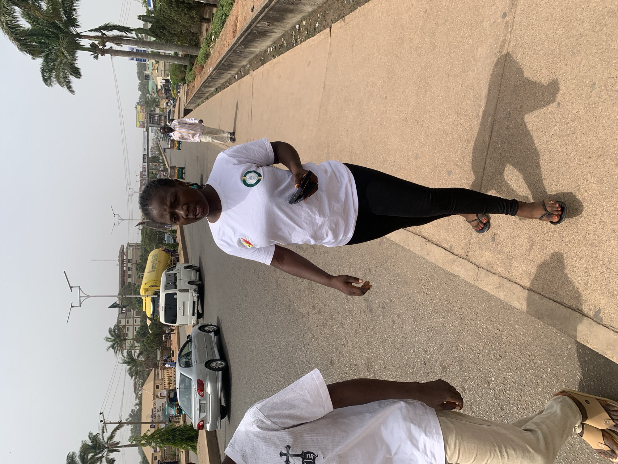 The image captures an outdoor urban scene in Jos, Nigeria, under bright, clear daylight, indicated by strong shadows. The primary subject is a young Black woman standing on a paved sidewalk, looking towards the camera with a neutral expression. She wears a white short-sleeved t-shirt with a circular green and white logo, black leggings, and black flip-flops with red-painted toenails. She holds a dark smartphone in her right hand.
In the foreground, the lower torso and legs of another person are visible, wearing a white t-shirt with a dark, stylized cross graphic, light-colored trousers, and brown sandals. Both individuals cast distinct shadows on the ground.
The background reveals a bustling street with several vehicles, including a silver sedan, two white minibuses, and a larger yellow vehicle with "Greenline" partially visible on its side. Palm trees and utility poles line the road. Further in the distance, buildings, billboards (one with "GLORY" and another possibly "SKY HOYO"), and other pedestrians are visible. The setting depicts a lively urban environment, common to Nigerian cities.