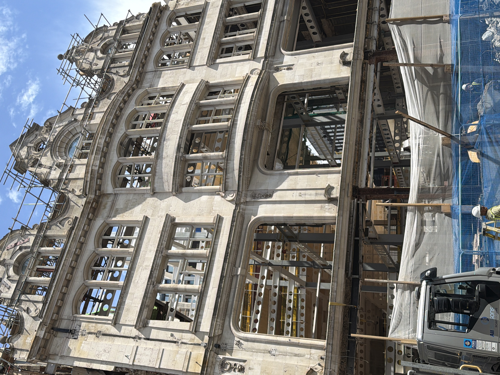 The image shows the former Midland Bank headquarters at 27-32 Poultry, London, now The Ned hotel, undergoing extensive renovations. The exterior of the building is visible, with scaffolding and protective sheeting partially obscuring the facade. The windows are either empty or reveal the building's internal steel structure.
A construction worker wearing a hard hat is partially visible at the right edge of the frame. Construction equipment, including a truck with "All Sons Scaffolding" printed on its side, is visible in the foreground. The sky is blue with scattered clouds in the background.