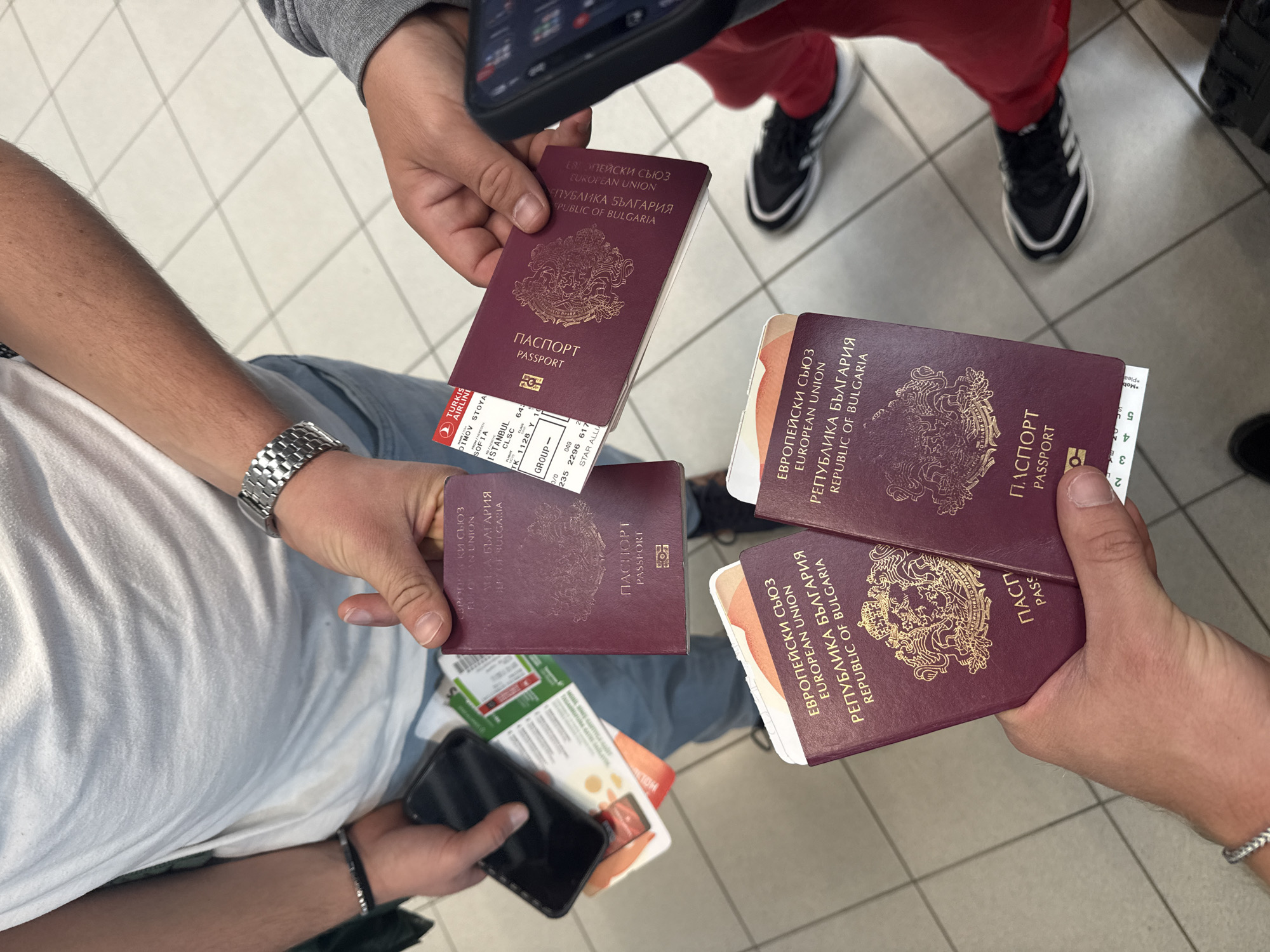 The image shows a group of four people standing on a tiled floor, likely in a public space such as an airport, given the context provided by the subjects and objects. Each person holds one or more Bulgarian passports. One person holds a Turkish Airlines boarding pass for a flight from Sofia to Istanbul with the passenger name Dimov Stoya. The location could be related to travel, and the boarding pass suggests it may be in Sofia, Bulgaria.
People: Four people are visible, though only their arms and hands are in focus. One person wears red pants and Adidas sneakers. Another person wears a white T-shirt.
Subjects: Bulgarian passports and a Turkish Airlines boarding pass.
Objects: Several Bulgarian passports, a Turkish Airlines boarding pass, a smartphone, a wristwatch, a black bracelet.
Actions: Holding passports and a boarding pass.
Interactions: The people are together, likely traveling as a group. They are displaying their passports and boarding pass.
Environment: The environment is a public space with a tiled floor. The context suggests the location is likely related to travel, possibly an airport.