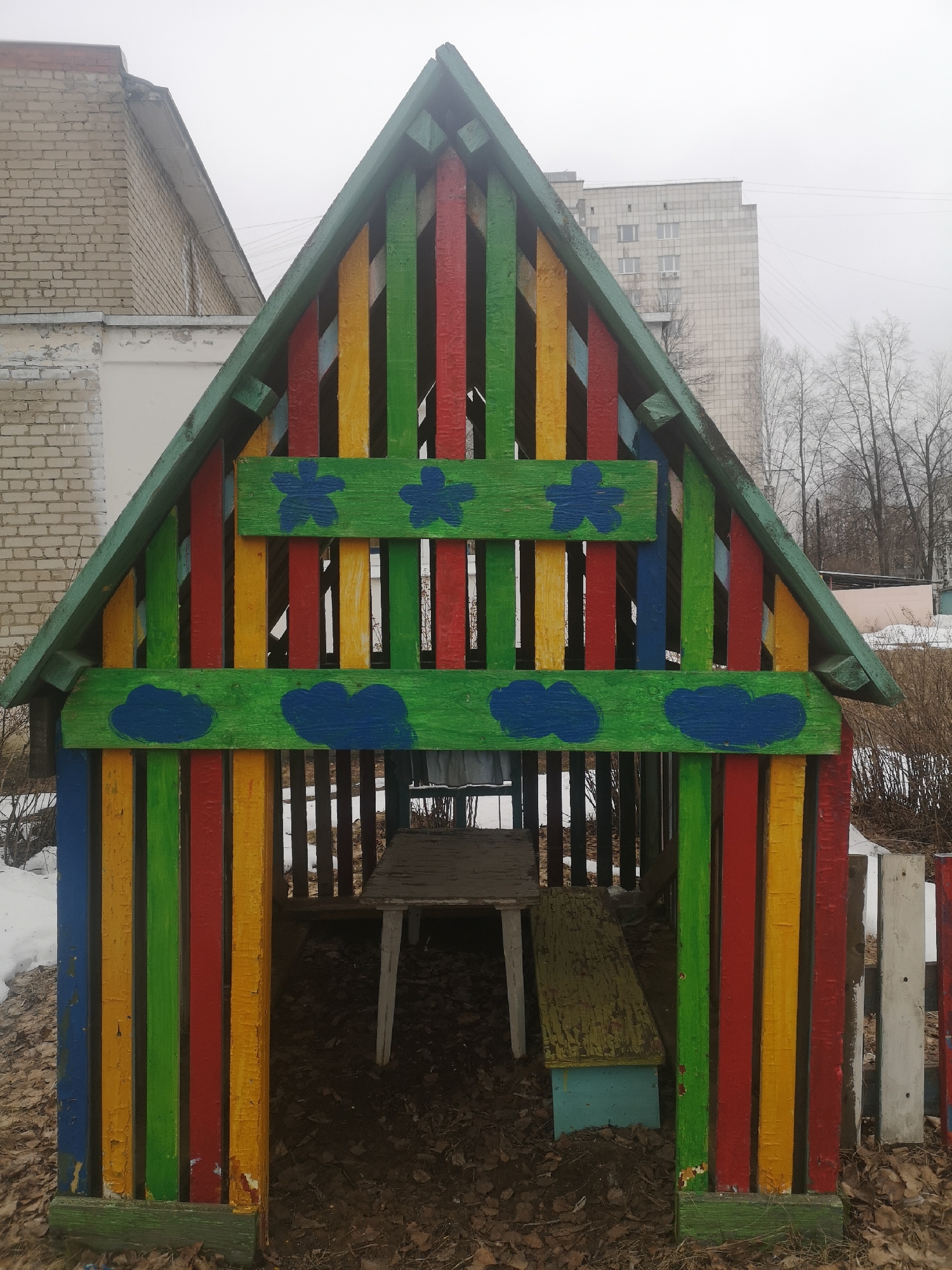 A small, colorful wooden playhouse stands outdoors, likely in Chaikovsky, Russia. The structure features vertical planks painted in alternating colors of blue, green, red, and yellow. The roof and horizontal beams are painted green, with blue flower and cloud designs. Inside the playhouse, a worn, light-colored table and bench are visible. The ground around the playhouse is covered in fallen leaves. In the background, there are buildings, trees, and remnants of snow. The sky appears overcast.