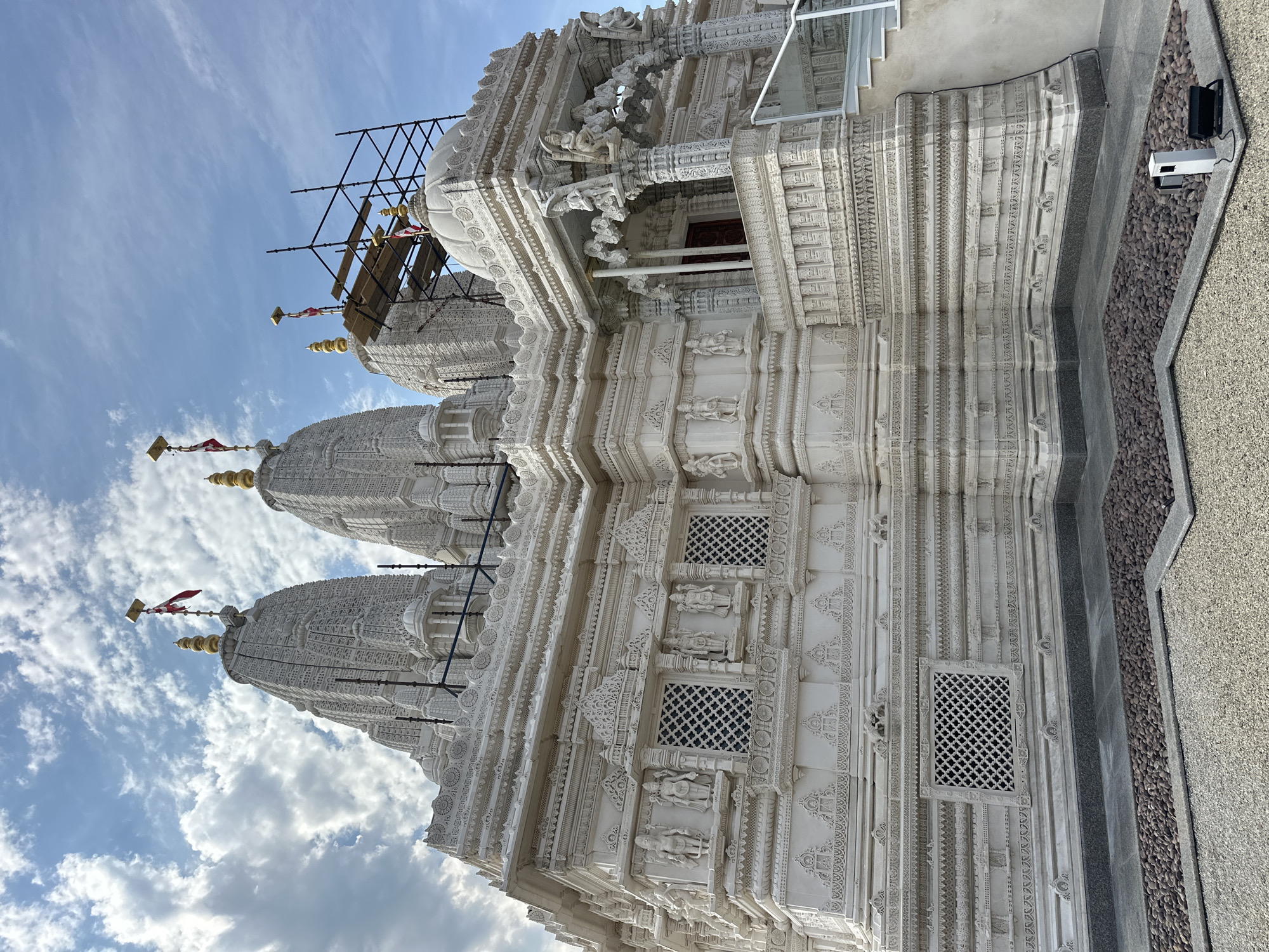The image shows a white marble temple with intricate carvings and multiple tiered roofs, some of which are adorned with golden finials. Scaffolding is visible around one of the roofs, suggesting ongoing construction or maintenance. Flags are positioned on some of the highest points of the temple. The temple is surrounded by a paved area with gravel and some decorative stones. In the foreground, there are two lighting fixtures. The sky above is partly cloudy. This is a view of the Shri Swaminarayan Mandir in Bartlett, Illinois, United States of America.